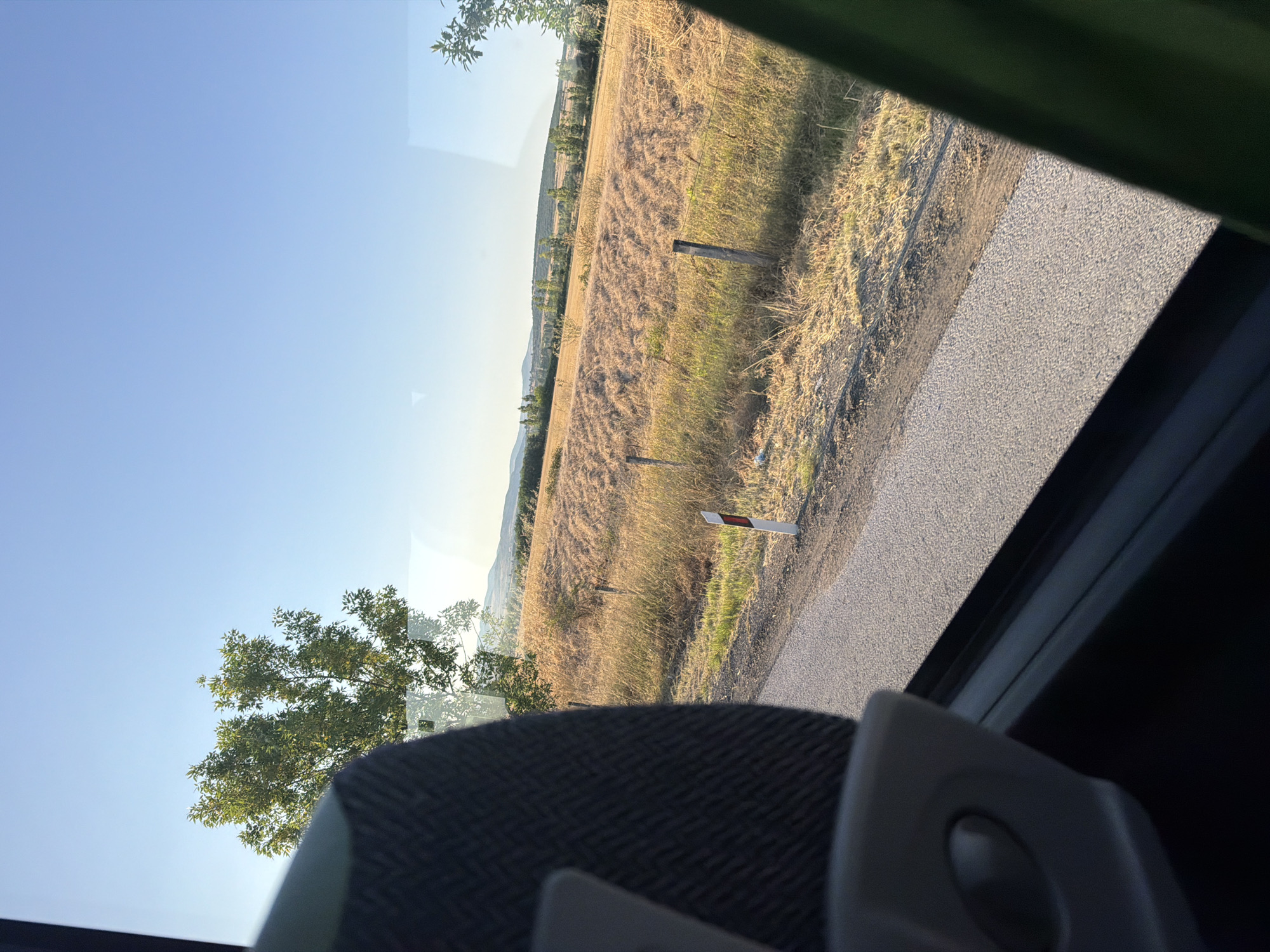 The image, taken from inside a vehicle, offers a view through the window of a rural landscape bathed in the warm light of what appears to be late afternoon. The foreground is dominated by the interior of the vehicle, with a portion of a dark fabric-covered seat on the left and a dark green curtain or fabric on the right. The window itself is visible, reflecting some elements of the interior.
Beyond the window, a paved road runs diagonally from the bottom left to the mid-right of the frame. Along the side of the road, a simple wooden fence runs horizontally. The fence posts are weathered and uneven. A white and red striped marker post stands near the edge of the road.
The landscape beyond the road consists of dry, golden-brown grass and fields, suggesting a dry or harvested season. Some areas show patterns of tilled earth or stubble. In the mid-ground, more trees are scattered, and a line of trees forms a distant horizon. The background is a clear, pale blue sky, with faint, hazy hills visible in the far distance.
The overall impression is one of a quiet journey through a rural area, with the sun casting long shadows and highlighting the textures of the dry vegetation and the road. The image does not contain any people or specific landmarks that would allow for definitive identification of the location as Shtarkovo, Bulgaria. However, the scenery is consistent with a rural setting in a region with similar terrain and vegetation.