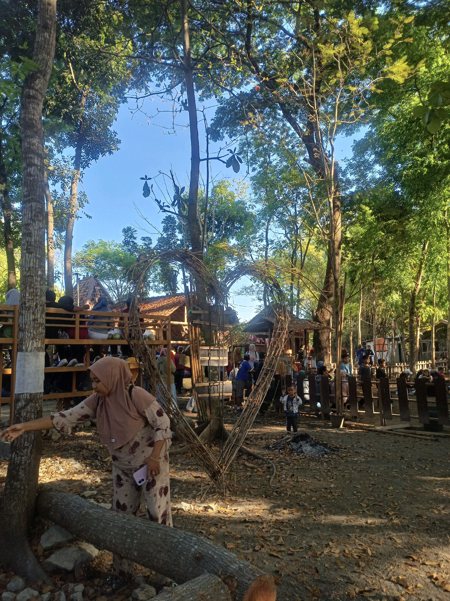 The image depicts an outdoor gathering place in the city of Karungan, Indonesia, characterized by a rustic, natural ambiance. The foreground is dominated by a woman in a light brown hijab and patterned outfit, reaching towards the base of a large tree. Behind her, a large, heart-shaped structure made of woven branches serves as a prominent visual element, with a wooden fence and more trees forming the background. Several small, traditional Indonesian-style structures with thatched roofs are visible behind the heart-shaped archway, suggesting a market or recreational area. People are scattered throughout the scene, some seated on wooden benches or standing in groups, interacting or observing their surroundings. The ground is covered with a layer of dry leaves and dirt, and scattered logs indicate a natural, possibly forested, environment. The presence of mature trees with lush green foliage suggests a daytime scene with ample natural light, casting dappled shadows across the ground. The overall atmosphere appears relaxed and informal.