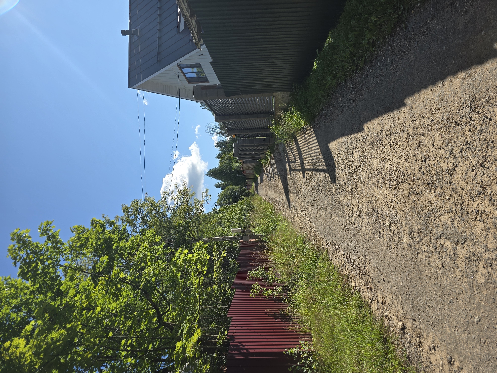 The image depicts a rural road in Lobkovo, Russia, on a clear, sunny day. A dirt path stretches into the distance, bordered on the left by a tall, corrugated red metal fence and lush green foliage. On the right, the path is lined with a dark green corrugated metal fence, behind which a modern-looking house with a dark gray metal roof and light-colored siding is visible. The house has a single visible window. Telephone wires are strung across the bright blue sky, which also features a few scattered white clouds. The sun casts strong shadows of the fence onto the dirt road. The environment is predominantly natural with man-made structures indicating a residential area.