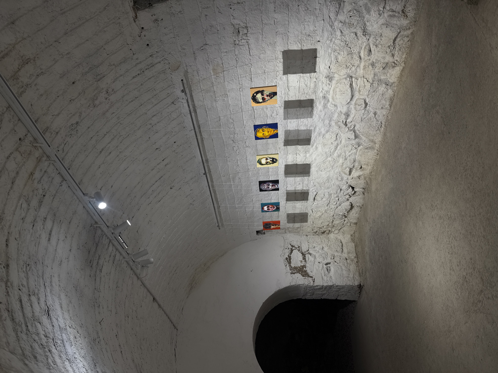 The image captures the interior of an arched space, likely a passage or tunnel, in what appears to be a structure with stone or brick walls, possibly in Yerevan, Armenia. The walls and arched ceiling are painted white, with visible texture and what seem to be aging marks on the surface. A series of small, colorful portrait images are hung in a vertical line, suspended by thin wires along one side of the arch. Shadows from these portraits are cast on the wall behind them. A track lighting system is mounted along the left side of the arched ceiling, with spotlights directed downwards. At the end of the arch, a dark opening suggests a doorway or continuation of the passage into darkness. The overall lighting in the scene is dim, emphasizing the contrast between the white surfaces and the darkness.