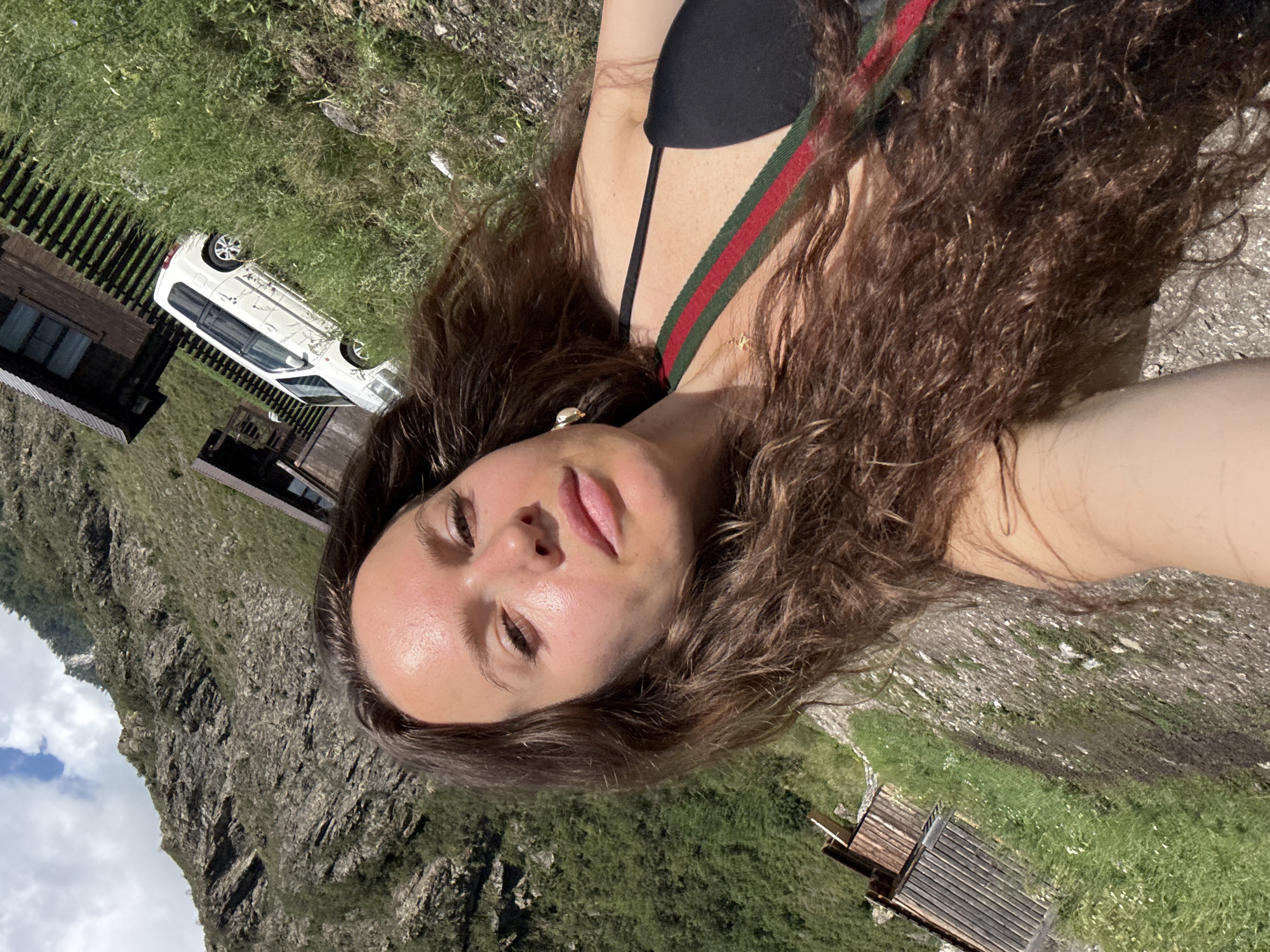 The image depicts a young woman in a close-up selfie, with her face and upper torso prominently featured. She has long, wavy brown hair that cascades around her shoulders and chest. She is wearing a black bikini top with a distinctive red and green striped strap. A delicate gold necklace with a small charm is visible around her neck. Her skin appears sun-kissed, with a few freckles visible on her chest. She is outdoors, and the background suggests a mountainous or hilly landscape with sparse vegetation and rocky terrain. Two wooden structures, possibly cabins or outbuildings, are visible in the distance, along with a white vehicle parked on a grassy incline. The sky is partially cloudy. The overall impression is a candid shot taken in a natural, possibly rural, setting. It is not possible to determine the exact location within Chemal, Russia, from the image alone.