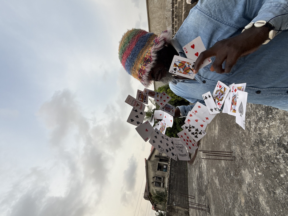 An adult male is depicted performing a playing card trick on a concrete rooftop in Ado, Nigeria, under an overcast sky. The man, with dark skin and a grey beard, wears a multi-colored striped and textured bucket hat with a furry brim, and a light blue denim-style button-up shirt. A cowrie shell bracelet is visible on his left wrist. Numerous playing cards are suspended in an arc-like formation to his left, appearing to defy gravity, with card faces like the 8 of Clubs, 7 of Diamonds, King of Diamonds, and Ace of Spades visible. Some cards bear text such as "LIONCOAL PLAYING CARDS." His right hand holds a Queen of Hearts and a Three of Hearts, with his right index finger extended towards the Queen. The rooftop ground is grey concrete, with several exposed rebar rods protruding from the surface on the left. In the background, a two-story building with a light-colored facade and distinct arched windows is visible, partially obscured by green foliage, next to a plain concrete wall.