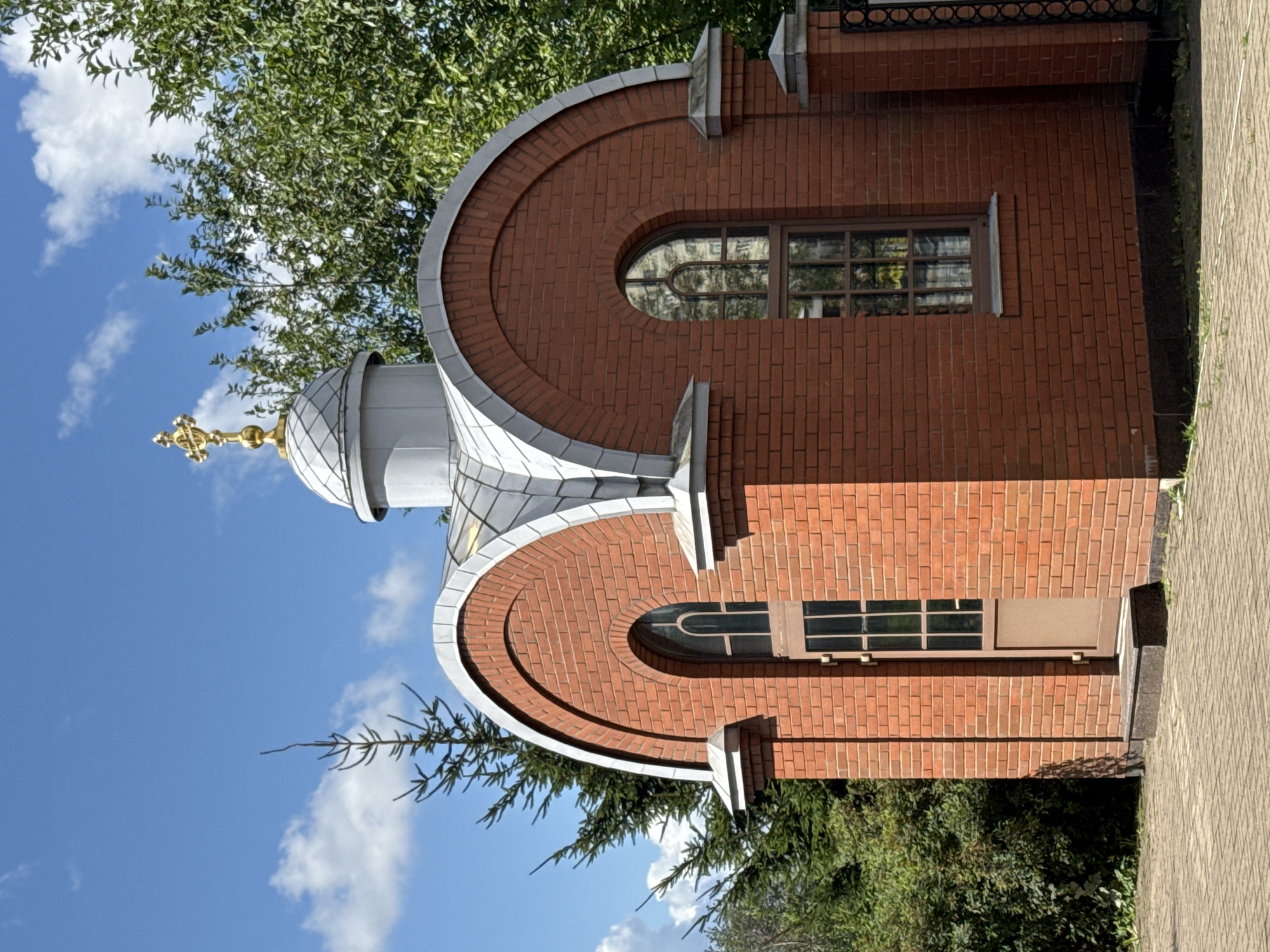 A small, red brick chapel with a silver domed roof topped by a golden cross stands under a bright blue sky with scattered white clouds. The chapel has arched windows with multiple panes and a single door. Green trees and foliage surround the structure, and a paved walkway is visible in the foreground. The chapel is located in Saint Petersburg, Russia.