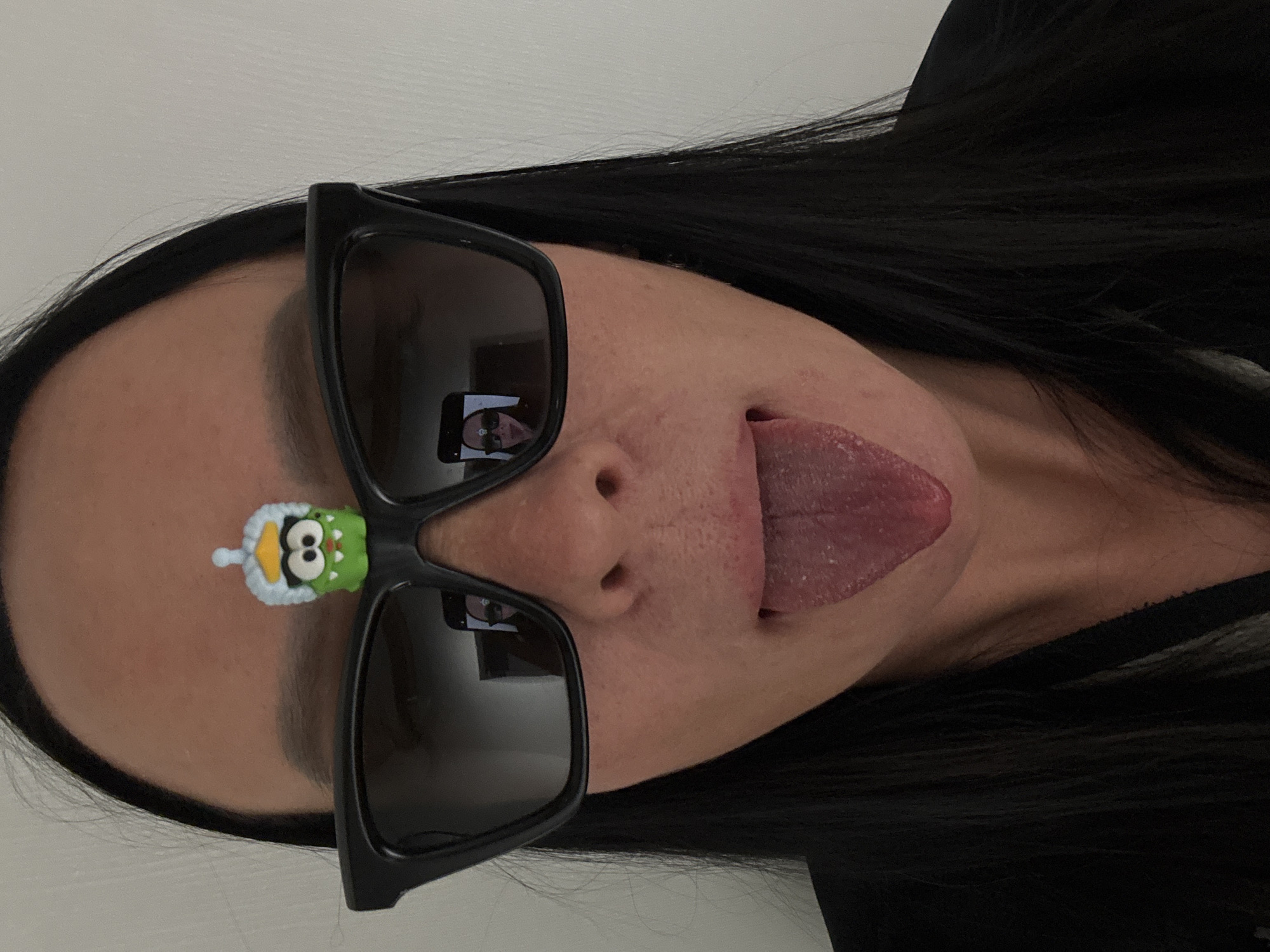 A woman with long, dark hair is pictured close-up. She is wearing black-framed sunglasses and sticking her tongue out. A green cartoon character is affixed to her forehead between her eyebrows. The reflection in the sunglasses shows a person holding a phone, likely taking the photo. The background appears to be a plain white wall. No information related to the city of Chaikovsky, Russia, can be determined from the image.
