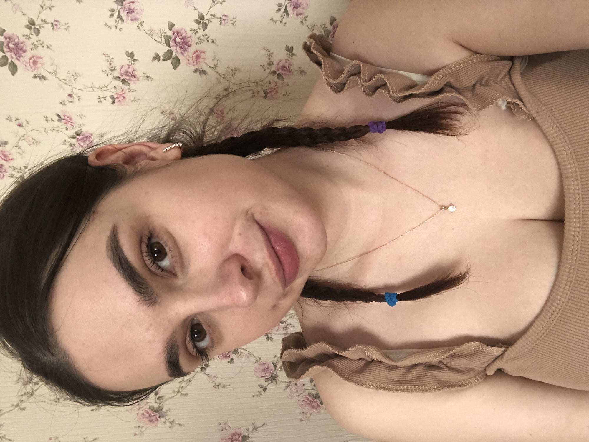 The image depicts a close-up, head-and-torso portrait of a single female subject in an interior setting. The visual content of the image provides no distinguishing features to confirm its specific geographic location as Noviy, Russia.
The subject is a light-skinned adult female with dark brown hair, parted centrally and styled into two braids that hang over her shoulders. The braid on the viewer's left is secured with a blue elastic, and the braid on the viewer's right is secured with a purple elastic. She has brown eyes, visible eyelashes, and well-defined eyebrows. Her lips are slightly parted in a faint smile, showing a natural pinkish hue, possibly enhanced with lip gloss. She wears small, multi-stone hoop earrings in both ears. Around her neck is a delicate gold-colored chain necklace with a single, clear, faceted stone pendant.
She is wearing a light tan or beige ribbed tank top or camisole with ruffled or wavy straps. A lighter-colored garment, possibly an undergarment, is visible beneath the top on her left side. The subject is looking directly into the camera.
The background consists of a wall covered with light-colored wallpaper, patterned with pink and purple roses and green foliage. The lighting appears even, suggesting an indoor light source. No other objects or environmental cues are visible.