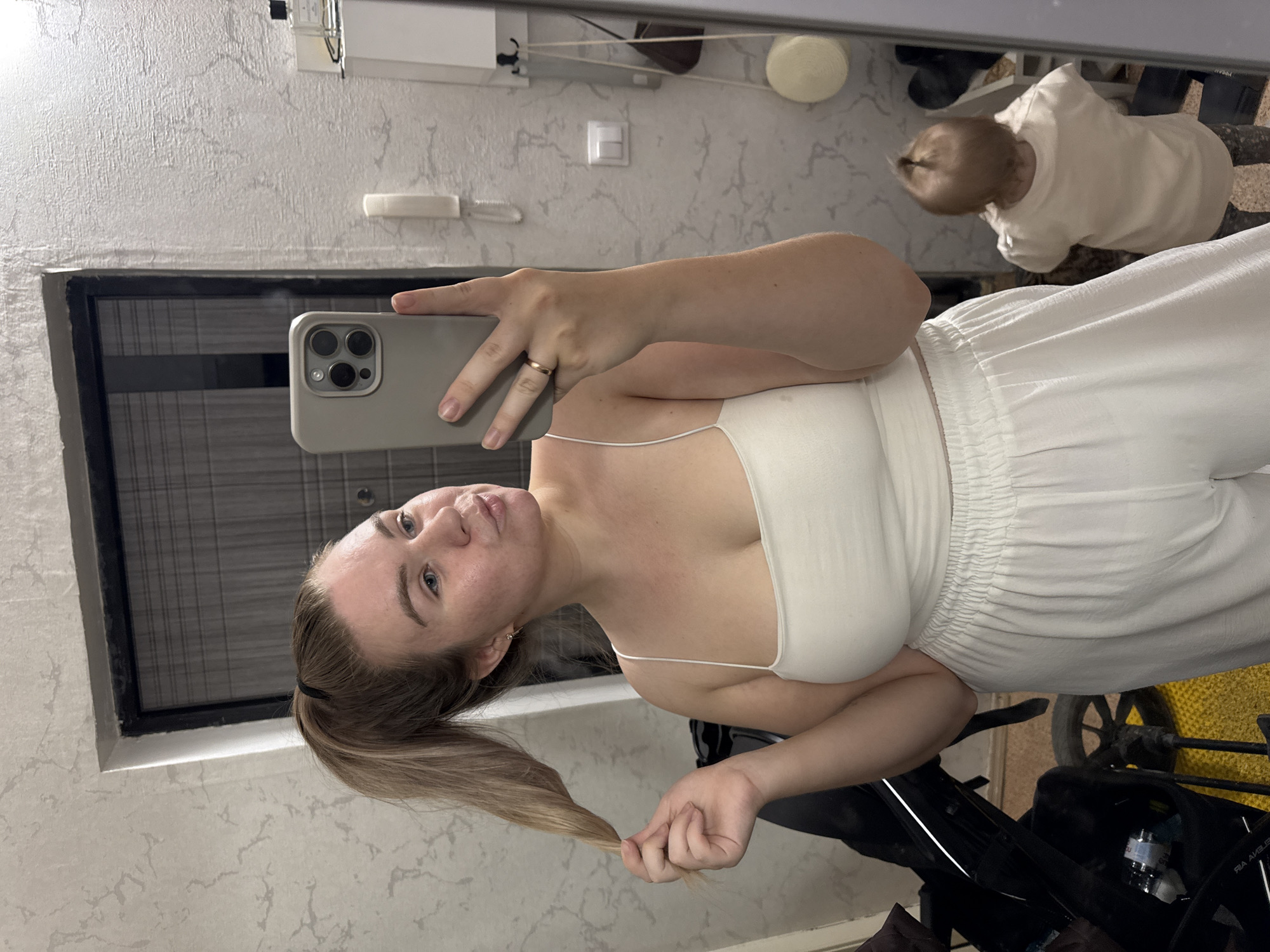 A woman takes a mirror selfie indoors. She has blue eyes and light brown hair pulled back in a ponytail. She wears a white spaghetti strap top and white pants. Her left hand holds the ends of her ponytail, while her right hand holds a smartphone with three cameras. The phone has a light grey case, and she wears a gold ring on her ring finger. Reflected in the mirror is a baby in a white shirt, a white wall, and some objects attached to the wall. A stroller is visible behind the woman. The location appears to be indoors in a home, possibly in Chaikovsky, Russia, based on contextual clues like architectural styles and common household items in that region.