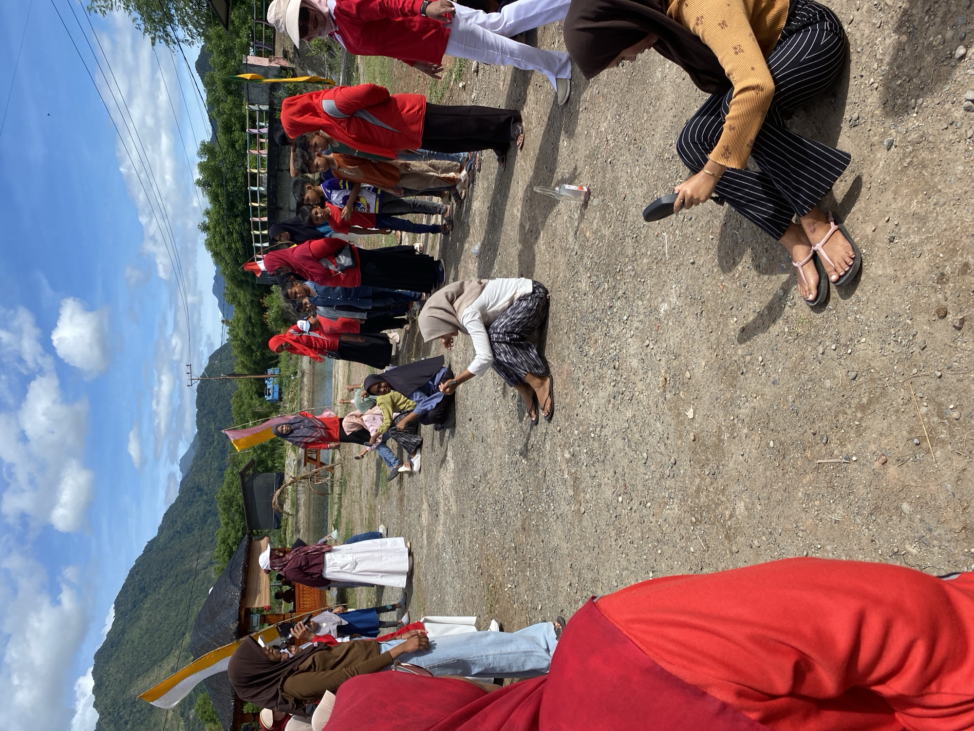 In Aneuk Paya, Indonesia, a group of people are gathered outdoors on a sunny day, with a backdrop of lush green mountains and a cloudy blue sky. The ground is covered in gravel and dirt.
In the foreground, a person wearing a red hooded jacket and blue jeans is partially visible on the left, facing away from the camera. To the right, another person in a brown headscarf and a mustard-colored top, with black and white striped pants and pink flip-flops, is sitting on the ground, holding a black object, possibly a phone or a small camera.
In the middle ground, several individuals are seated on the ground. One person wearing a hijab and a light-colored top with patterned pants is also sitting. Further back, a group of children and adults stand in a line. Some of the adults are wearing red, and one woman is holding a white flag with red and white stripes. Another woman in a red headscarf stands with a group of children in red shirts. A few people are seen near a small, open-air shelter with a thatched roof.
Objects in the scene include flags, a bottle on the ground, a bicycle in the background, and power lines overhead. The overall atmosphere appears to be a community gathering or event.