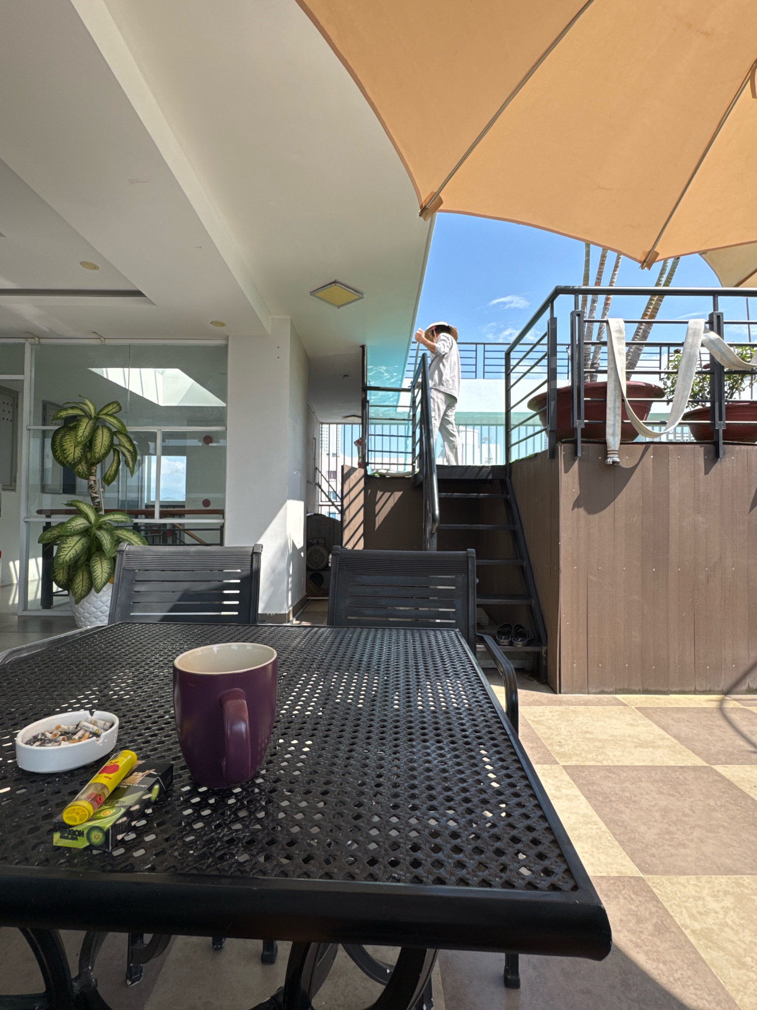 The image depicts an outdoor seating area with a black metal table and chairs. On the table, there is a purple mug, an ashtray with cigarette butts, and a pack of cigarettes with a lighter. In the background, a person wearing a straw hat and light-colored clothing is standing on a balcony, looking out. A set of stairs leads up to the balcony, and there are potted plants and a large umbrella providing shade. The scene appears to be from a rooftop cafe or restaurant in Nha Trang, Vietnam, given the tropical plants and the bright, sunny weather. The tiled floor with a checkered pattern suggests a communal outdoor space.