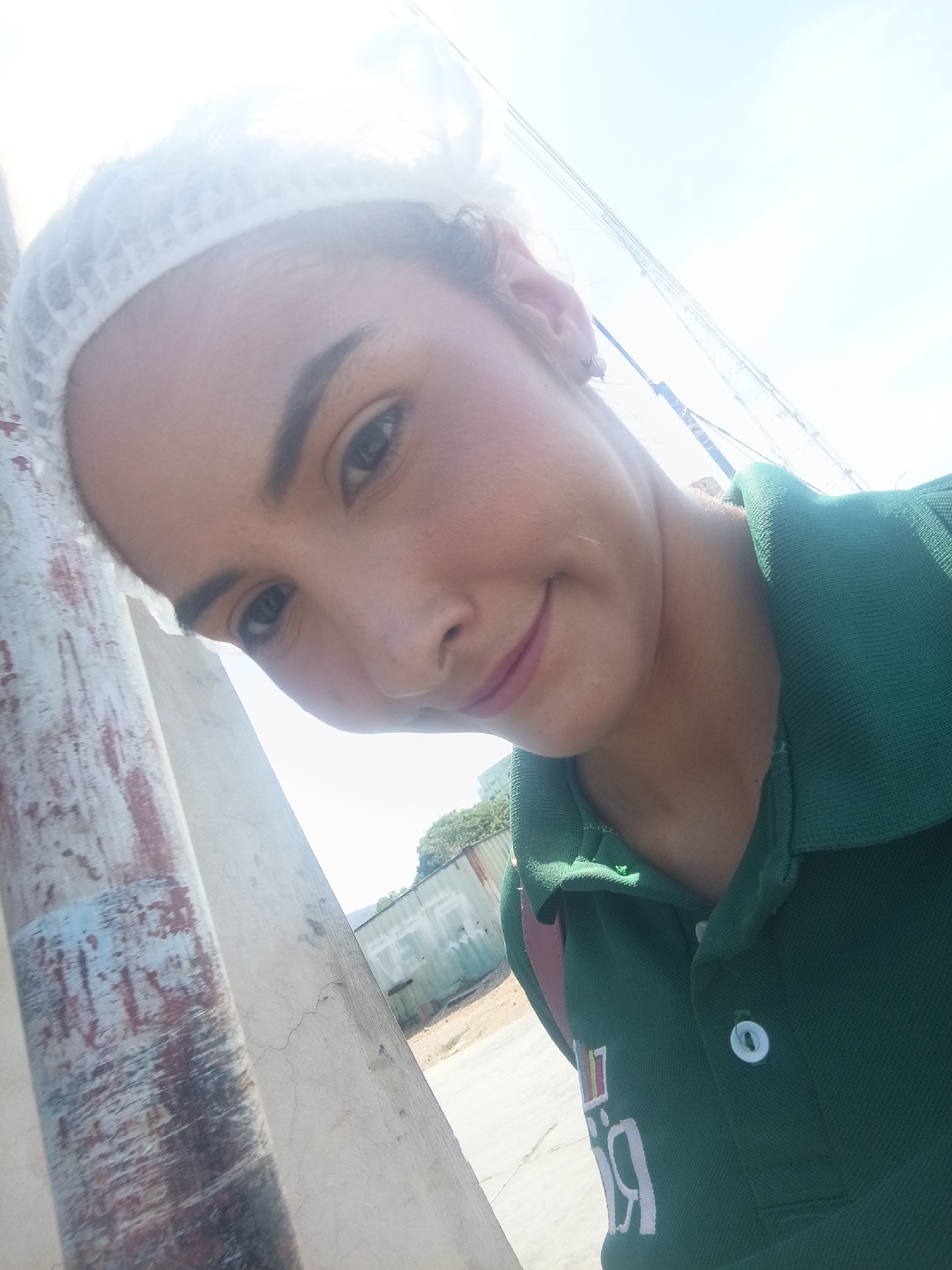 A woman in a green polo shirt and a white hairnet is posing for a selfie. She is leaning against a concrete pillar that is painted with peeling gray, brown, and blue paint. Behind her, there is a corrugated metal fence and some vegetation. The sky is bright blue. It is likely that the location is in the city of Juan Griego, Venezuela, as stated in the instructions.