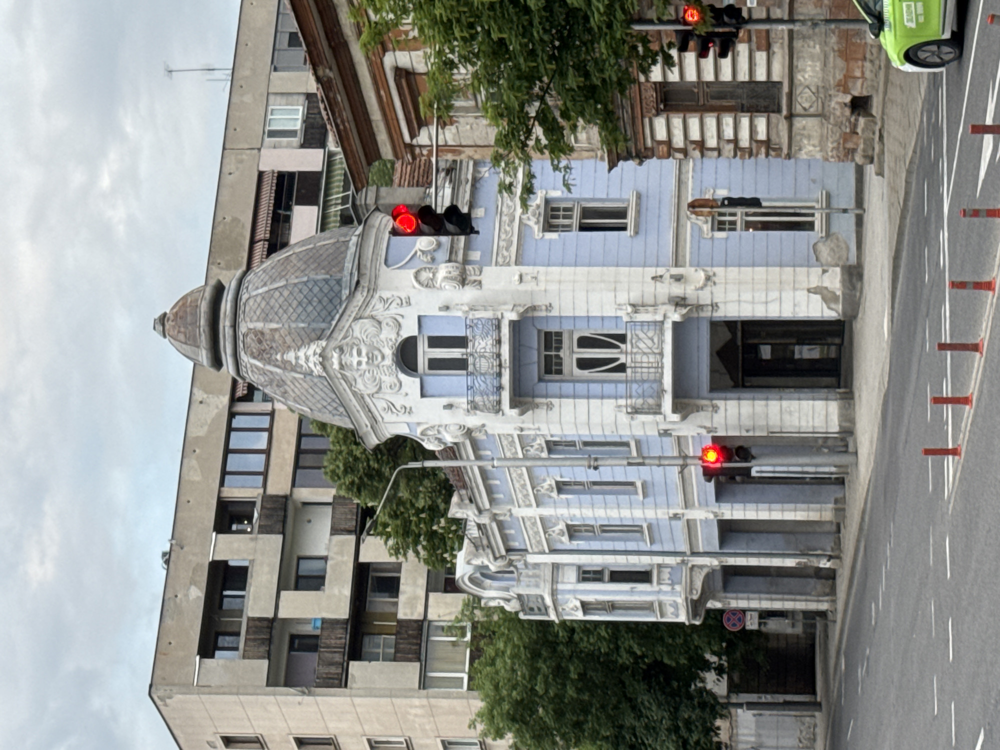 A daytime, eye-level shot shows a building and street scene in Plovdiv, Bulgaria. The image is rotated counterclockwise about 90 degrees.
The focus is on an ornate building with a grey dome and light blue and white facade. The building is adjacent to a plain, taller, multi-story building. The street is visible to the right with road markings and red lane dividers. The traffic lights are red. A green car is partly visible at the right upper corner. Trees are visible throughout the shot, obscuring parts of the buildings.