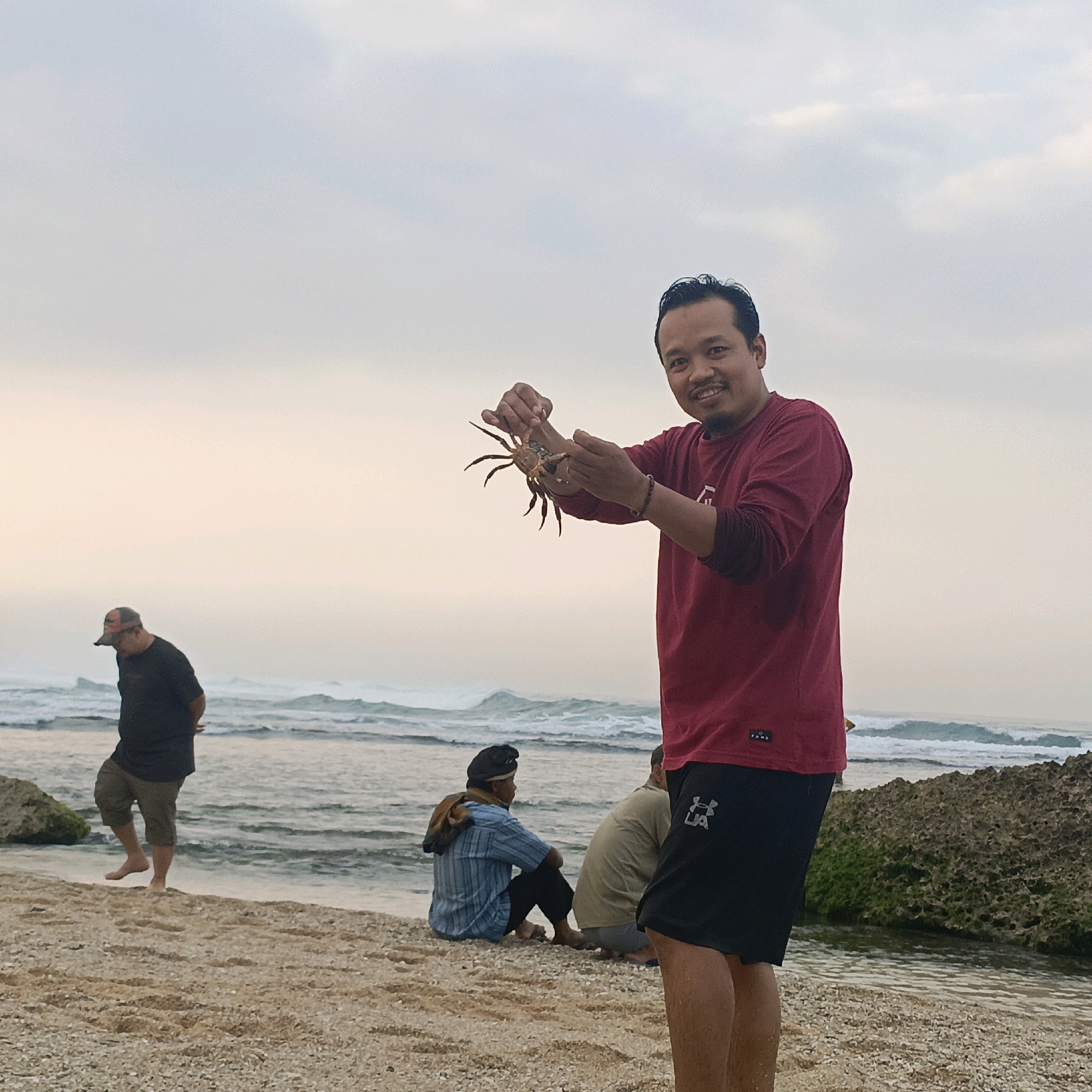 In Tepus, Indonesia, a man stands on a beach, holding a crab. He is smiling and looking towards the camera. The man is wearing a red long-sleeved shirt and black shorts. Behind him, the ocean stretches out to the horizon, with waves gently crashing on the shore. Several other people are visible on the beach, some sitting and others walking. The sky is overcast, with a soft, diffused light. The beach itself is sandy, with some rocks and seaweed scattered along the waterline. The overall atmosphere is relaxed and peaceful.