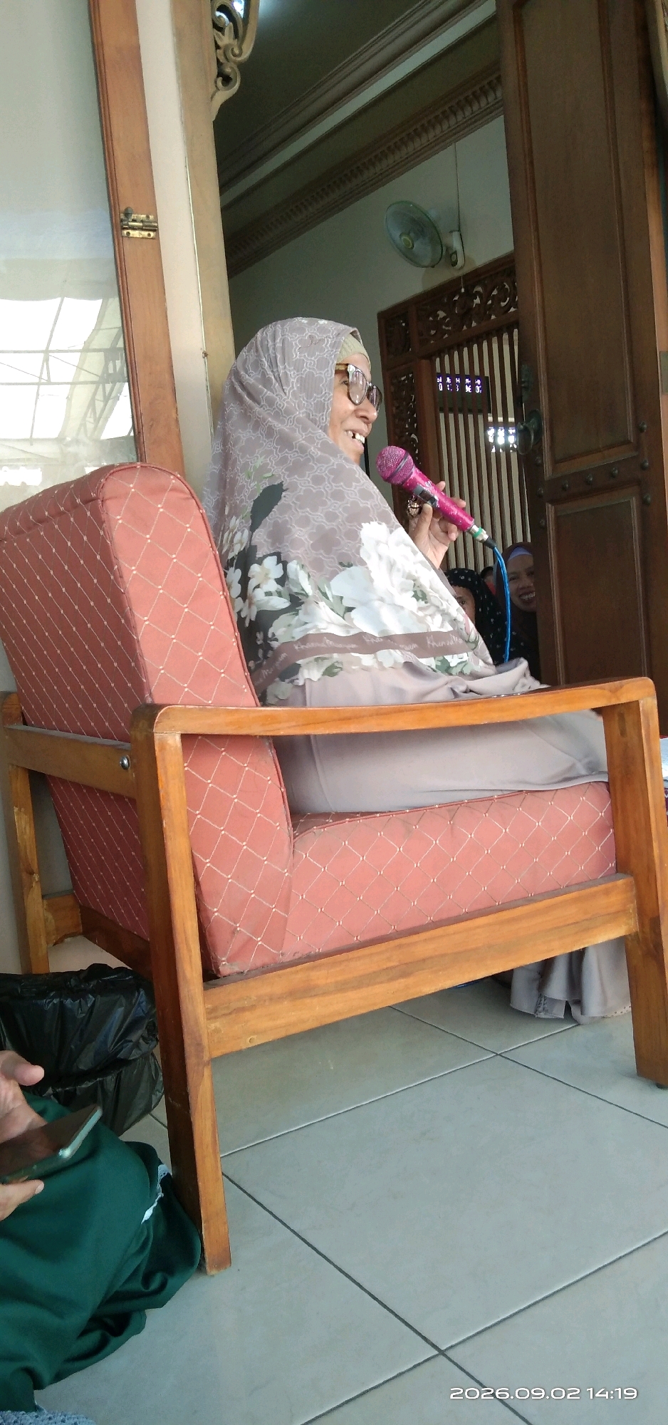 An elderly woman wearing a light brown hijab and glasses is seated in a cushioned wooden chair, holding a pink microphone. She appears to be speaking or singing, with her mouth open. The chair has a distinctive diamond pattern on its fabric. In the background, another person with a dark head covering is visible, smiling and peeking from behind a doorway with ornate wooden carvings. The scene takes place indoors, with a tiled floor and a window with a wooden frame visible to the left. A black trash bag is on the floor near the chair. The image was taken on September 2, 2026, at 2:14 PM, and is a photograph from Panembahan, Indonesia.