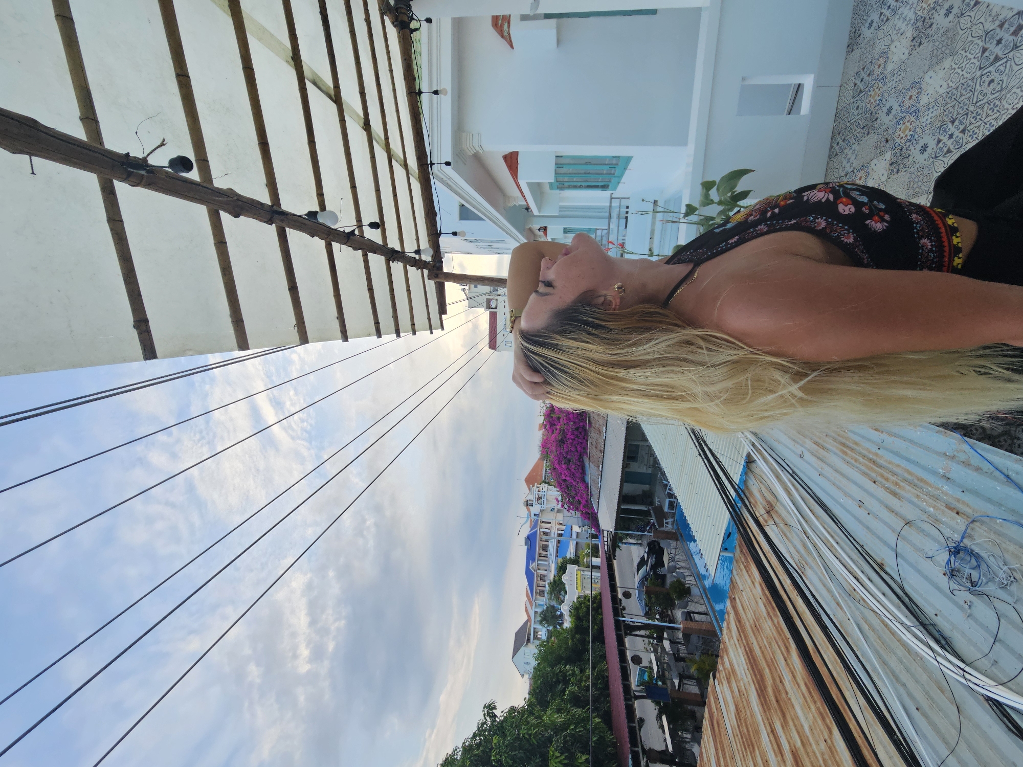 This media file features a woman with long blonde hair lying down outdoors. She is wearing a dark, floral-patterned top and a necklace. Her head is resting on her hand, and her eyes are closed, suggesting a relaxed or contented state.
The setting appears to be a rooftop or balcony, with a bamboo-covered awning overhead and strings of lights. In the background, a colorful urban landscape unfolds, with buildings of various colors, including white, blue, and red. Lush green trees and vibrant pink flowers add to the scene. The sky is visible, with scattered clouds indicating it might be late afternoon or early evening.
The overall atmosphere is tranquil and leisurely, as if the woman is enjoying a moment of repose in a pleasant outdoor environment. The presence of buildings and overhead wires suggests an inhabited area, possibly a town or city. The scene seems to be set in Phu Thuy Ward, Vietnam, as indicated by the location context. There is no visible text in the image.