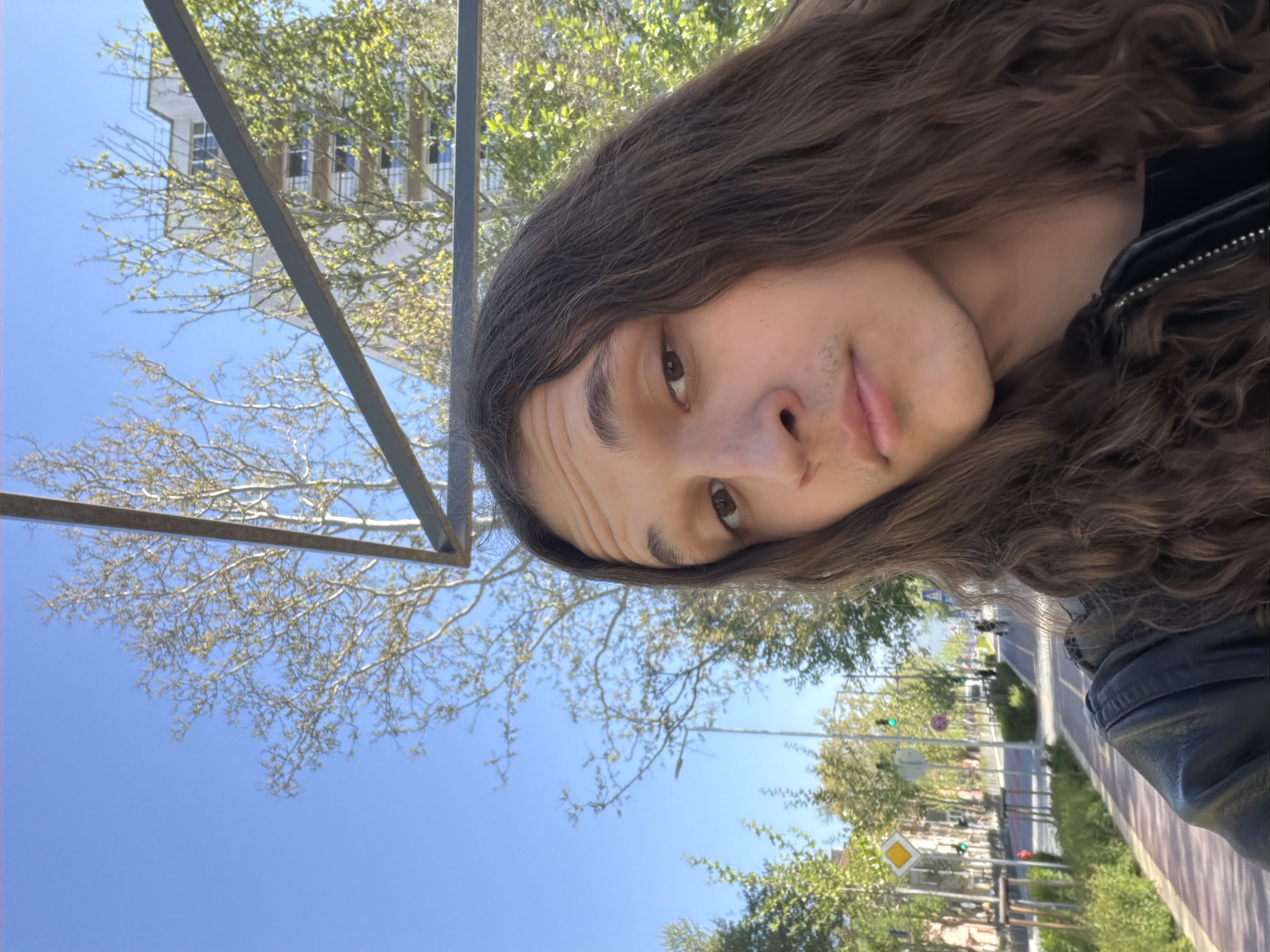 This image is a brightly lit, rotated outdoor selfie. The main subject, a young person with long, wavy brown hair, occupies the right side of the frame, their head sharply tilted to the right. Their right eye, nose, and mouth are visible, featuring light stubble on their upper lip and chin, and a neutral expression. They are wearing a dark, possibly leather, jacket with a visible zipper detail on the shoulder. Their hair flows towards the bottom of the image.
The background is an urban or suburban setting under a clear, bright blue sky. A dark metal bar or frame crosses diagonally through the upper center of the image. Through and above this, bare tree branches with emerging green leaves (indicating spring) are visible. In the upper left, a white multi-story building with dark windows can be seen.
The lower left and bottom portions of the image reveal a street or pathway, with yellow and white road markings. Traffic lights (one green, one red in the distance) and a distinctive yellow diamond-shaped road sign, common for indicating a priority road in Europe, are discernible. More green trees line the street. The bright natural sunlight indicates it is daytime. No specific text is clearly legible in the image. The scene suggests a sunny day in a European urban environment, consistent with the Plovdiv, Bulgaria location context.