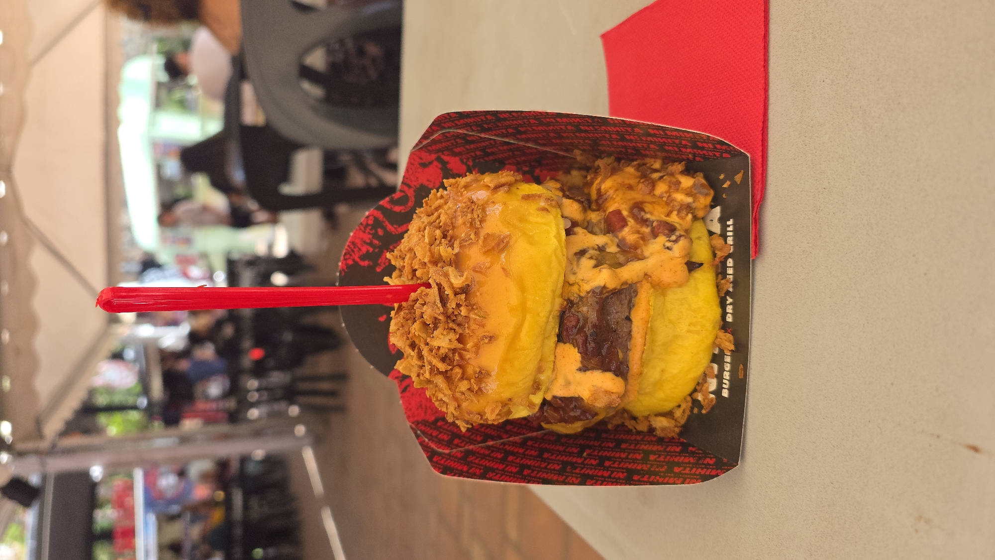 This is a close-up, slightly tilted, eye-level, outdoor shot of a burger served in a black and red paper container, possibly from a food stall or casual eatery, with the text "BURGER DRY AGED GRILL" visible on the container's edge. The burger is held together by a red plastic skewer and is topped with what appear to be crispy fried onions and a generous drizzle of orange sauce, possibly cheese sauce or a spicy aioli. The bun is golden yellow and appears soft.
In the background, the scene is blurred, suggesting an outdoor setting, likely a public space like a street market or a plaza, given the presence of other people and what looks like white tenting or awnings, common in outdoor events or temporary food setups. The lighting suggests it's daytime, possibly late morning or afternoon, with diffused light. The overall ambiance is casual and focused on the food. The vibrant red of the skewer and the napkin peeking out from the right add pops of color to the image. The visible text on the packaging indicates the burger is from a place that specializes in dry-aged burgers.