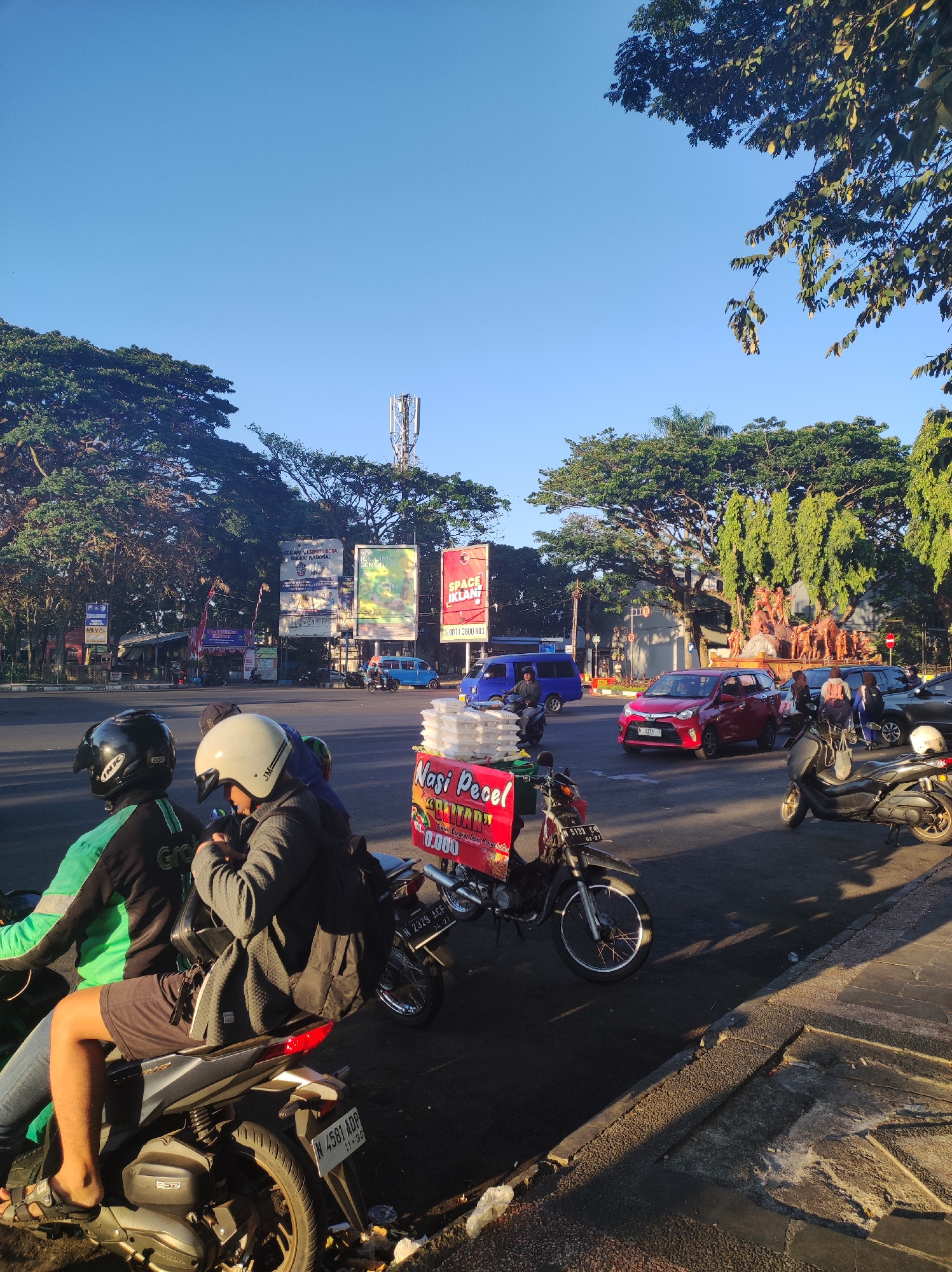 The image depicts a street scene in Malang, Indonesia, likely during the early morning due to the low sun angle and long shadows. In the foreground, two people are seated on a motorcycle, facing away from the camera. The person in the back wears a white helmet and a dark jacket, while the person in front wears a black helmet and a green jacket with the "Grab" logo visible. Adjacent to them is a food cart selling "Nasi Pecel," a traditional Indonesian dish, with stacks of white food containers.
The background reveals a busy intersection with several vehicles, including red cars, a blue van, and other motorcycles. Traffic lights and street signs are visible, along with advertisements displayed on large billboards. Towering trees line the sides of the road, and a cell tower is prominent in the distance. A large public sculpture, depicting several figures, is situated at the center of the intersection, suggesting a significant landmark within the city. The overall atmosphere is that of a typical urban environment in Indonesia during the daytime.