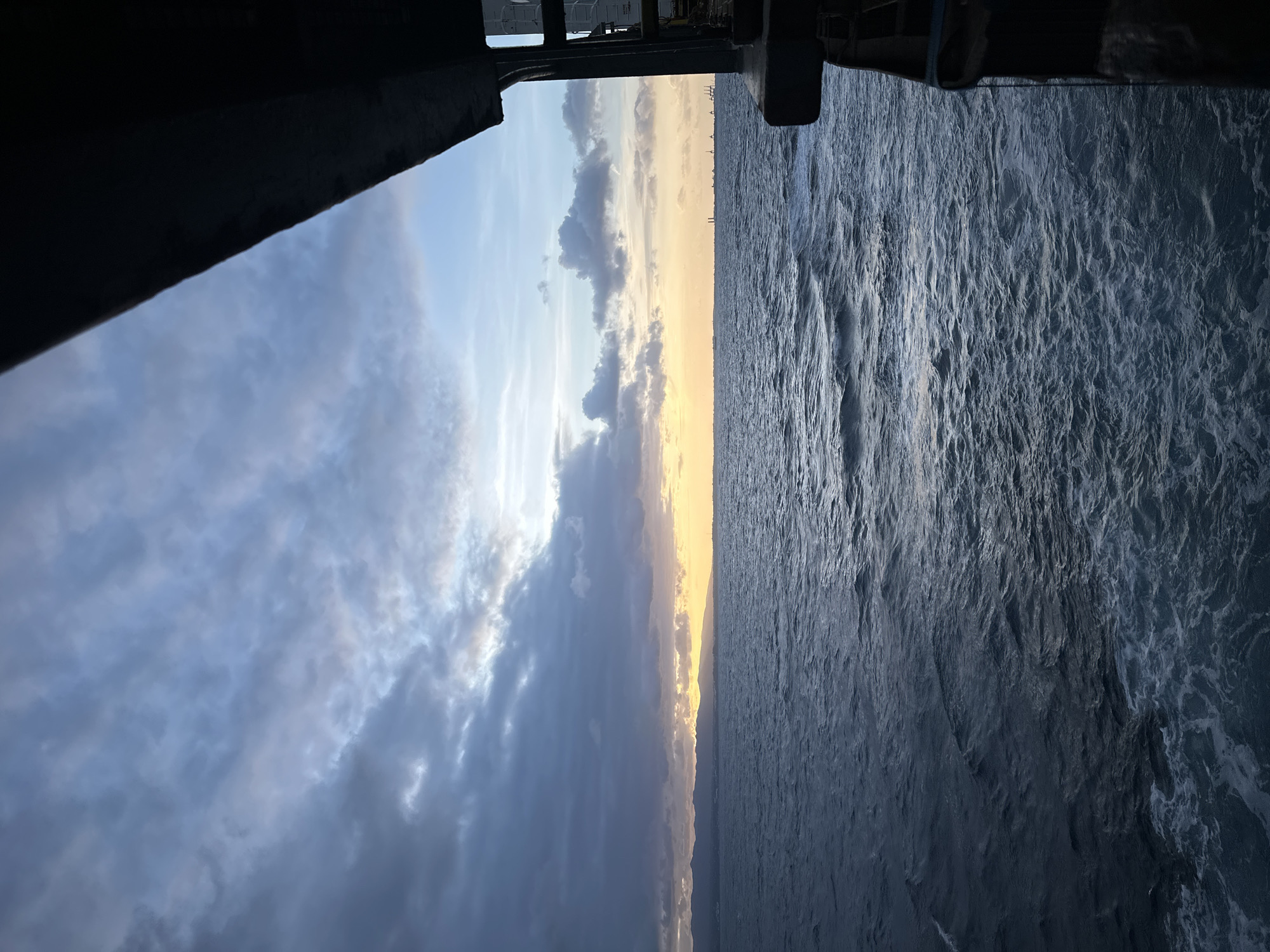 This image depicts a vast expanse of the ocean under a cloudy sky, likely taken from the deck of a vessel. The water is a deep blue, with visible waves and white foam churning on the surface. On the horizon, a faint line of land with mountains is visible, bathed in the warm glow of a sunset or sunrise. The sky is filled with dramatic clouds, varying in color from dark grays to lighter blues and hints of orange and yellow from the setting or rising sun. To the right, the dark silhouette of the vessel's structure intrudes into the frame, suggesting a perspective from a ship or ferry. There are no people visible in the scene, and the focus is on the natural elements of the sea and sky, as well as the imposing structure of the vessel. The overall atmosphere is serene yet dramatic, with the interplay of light and shadow across the clouds and water. The location in the city of Le Gosier, Guadeloupe, is not discernible from the visual elements present in the image.