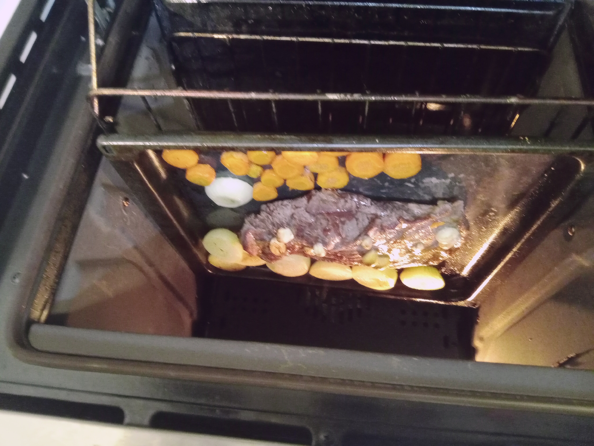 The image shows a meat roast with sliced carrots, sliced potatoes, and onion roasting in a black pan inside an oven. The roast appears to be cooked through. The location is Unknown, Unknown.