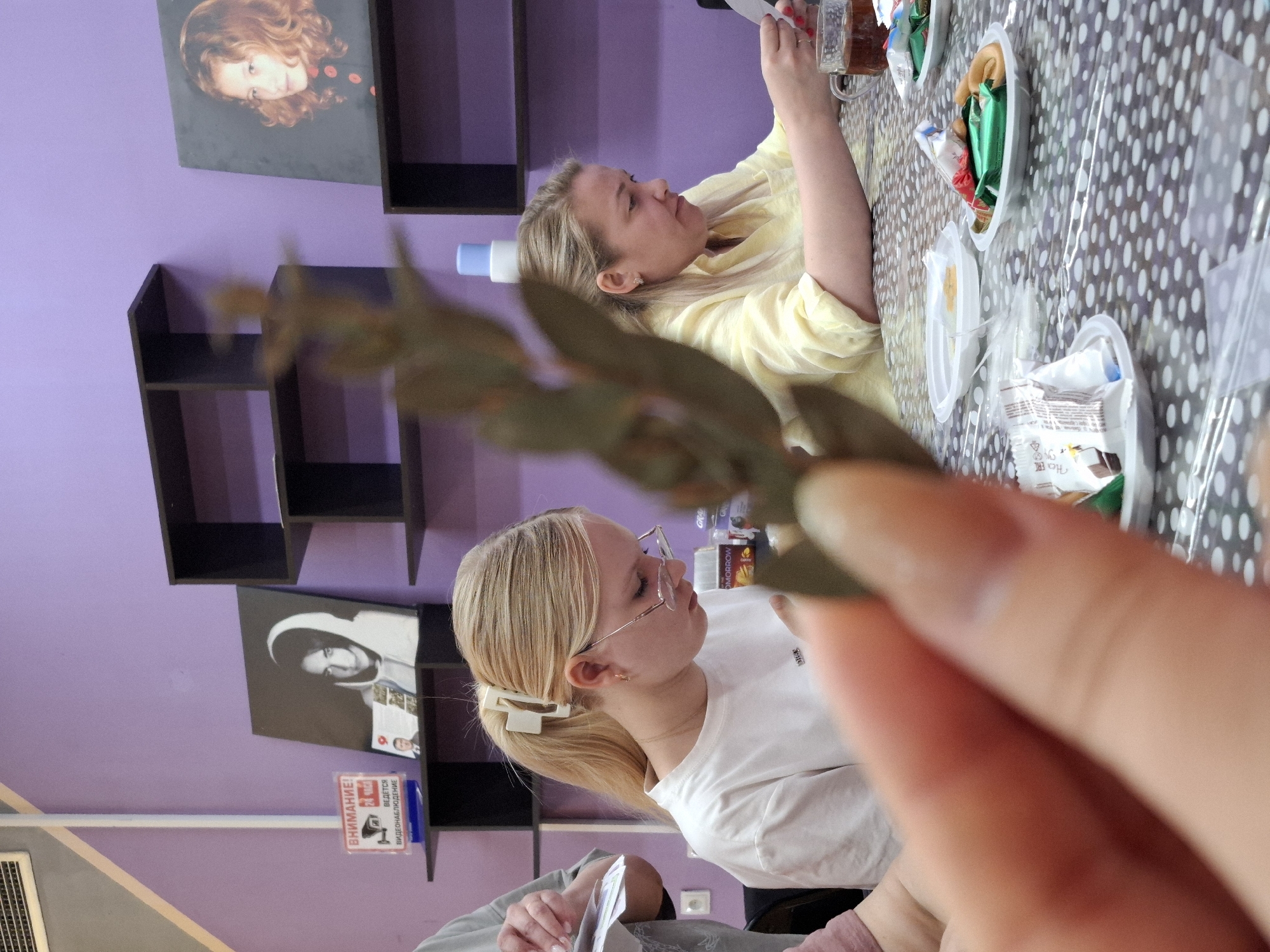Two fair-skinned women are seated at a table laden with food items in disposable plates. One woman is wearing a yellow shirt, and the other is in a white shirt, with blonde hair pulled back in a ponytail and wearing glasses. The background is a lavender wall with black shelving units and two framed images. A sign in Russian is visible, along with a glimpse of an air conditioning unit. A hand holding greenery is in the foreground, partially obscuring the view. The location is potentially Markovskii, Russia.