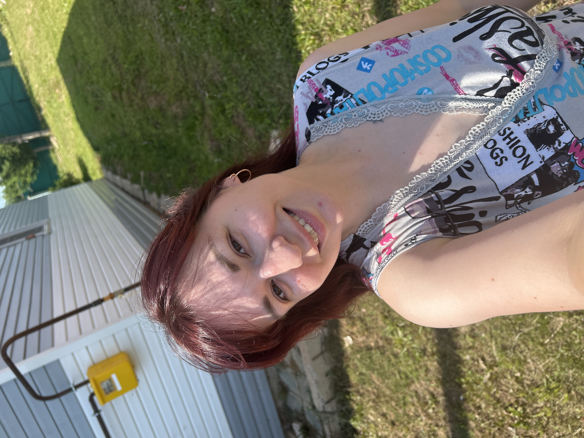 A woman with auburn hair is taking a selfie outdoors, smiling at the camera. She is wearing a grey tank top with a repeating pattern of text and images, including the words "BLOGS" and "COSMOPOLITAN". A gold hoop earring is visible in her left ear. Behind her, a light-colored house with grey siding is visible. A yellow gas meter is mounted on the side of the house, connected to a dark pipe. The ground in front of the house is covered in grass, with a shadow cast from an unseen object. The background shows more green grass and trees, with a teal fence. The overall setting suggests a suburban or rural environment, possibly in the city of Foky, Russia.