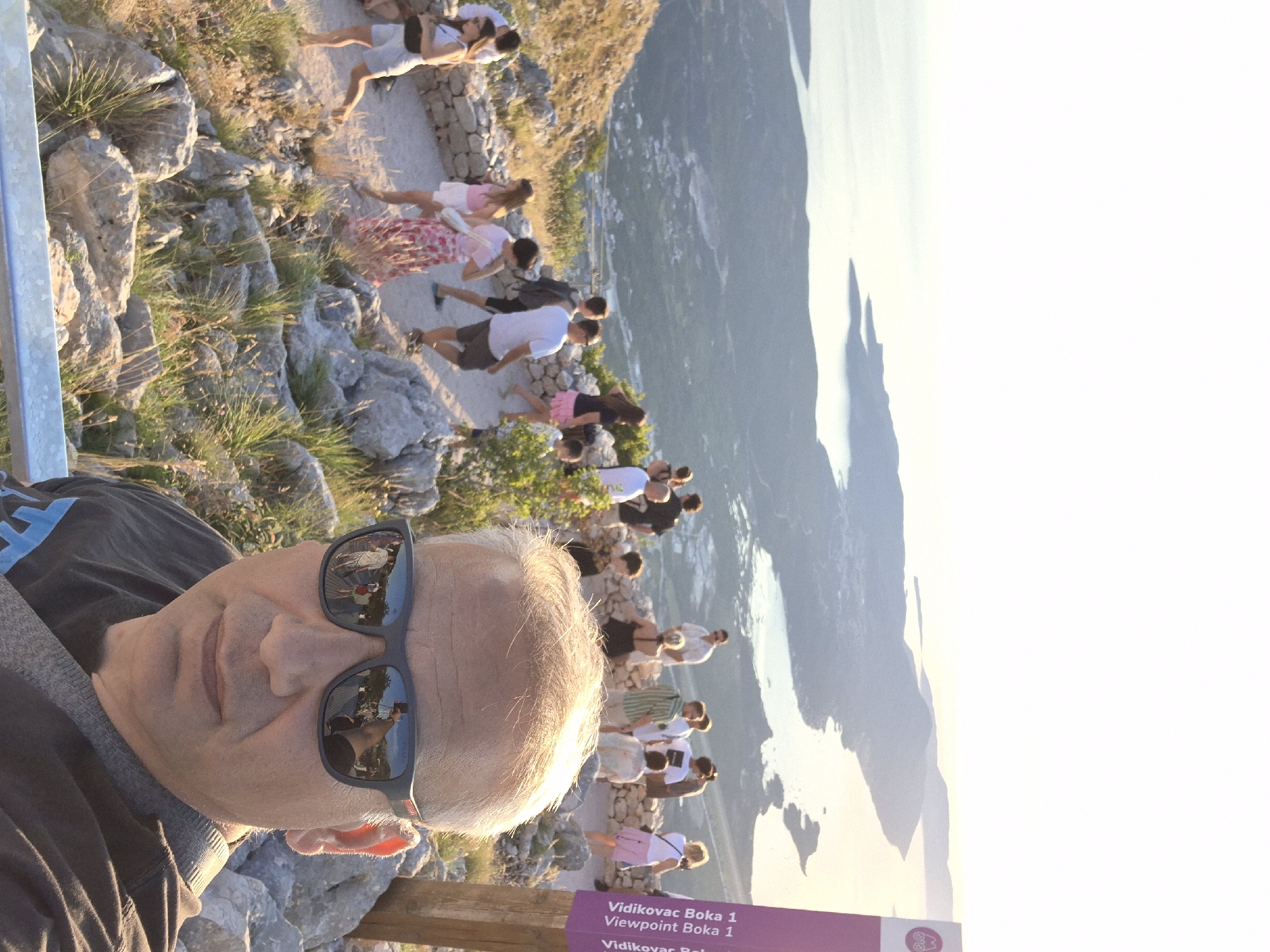 In the foreground, a man wearing sunglasses takes a selfie, his reflection showing a group of tourists and a scenic viewpoint. In the background, a diverse group of people are gathered on a stone pathway, overlooking a breathtaking coastal landscape of mountains and the sea. The location is identified as Vidikovac Boka 1, a viewpoint in the city of Kuk, Montenegro. The sky is clear and the sun is low in the sky, casting a warm glow over the scene.