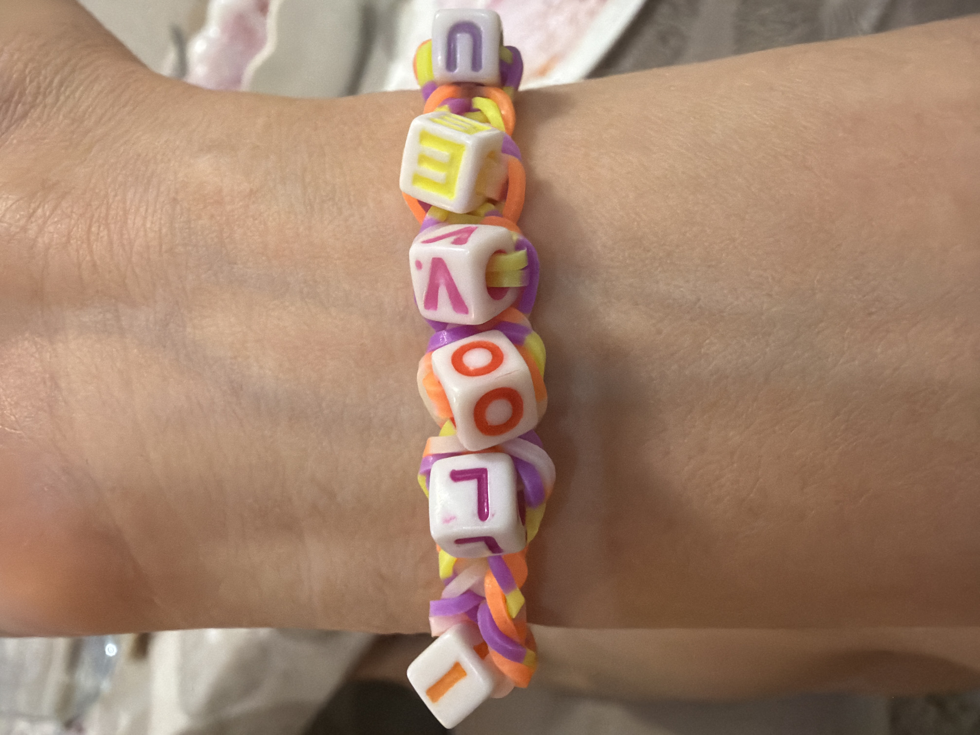 A close-up shot shows a person's arm with a colorful handmade bracelet. The bracelet is made of rubber bands and square letter beads spelling out a word or phrase. The rubber bands are arranged in a looped pattern, and the beads are strung along the loops. The colors of the rubber bands include yellow, orange, and purple. The letter beads are white with different colored letters, including purple, yellow, and orange. The arm is light-skinned, and the bracelet is positioned on the wrist. The background is slightly blurred and shows a white surface with indistinct objects. The image focuses primarily on the bracelet and the arm wearing it.
There is no information available to determine the location of the image in Budva, Montenegro, or if the image was taken there.