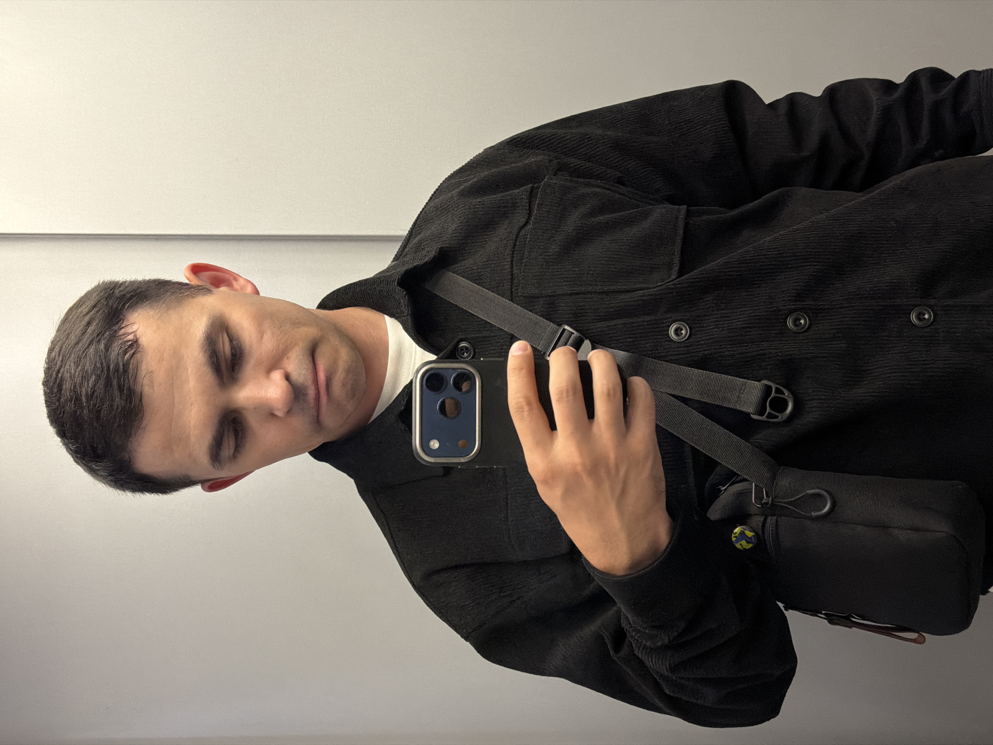 A man is holding a black phone in his hand, obscuring his face, which is turned slightly downward. He is wearing a dark, corduroy button-up shirt over a white t-shirt. A black strap crosses his torso, attached to a small, dark bag. The background is a plain, light-colored wall. There is no indication of Izhevsk, Russia.