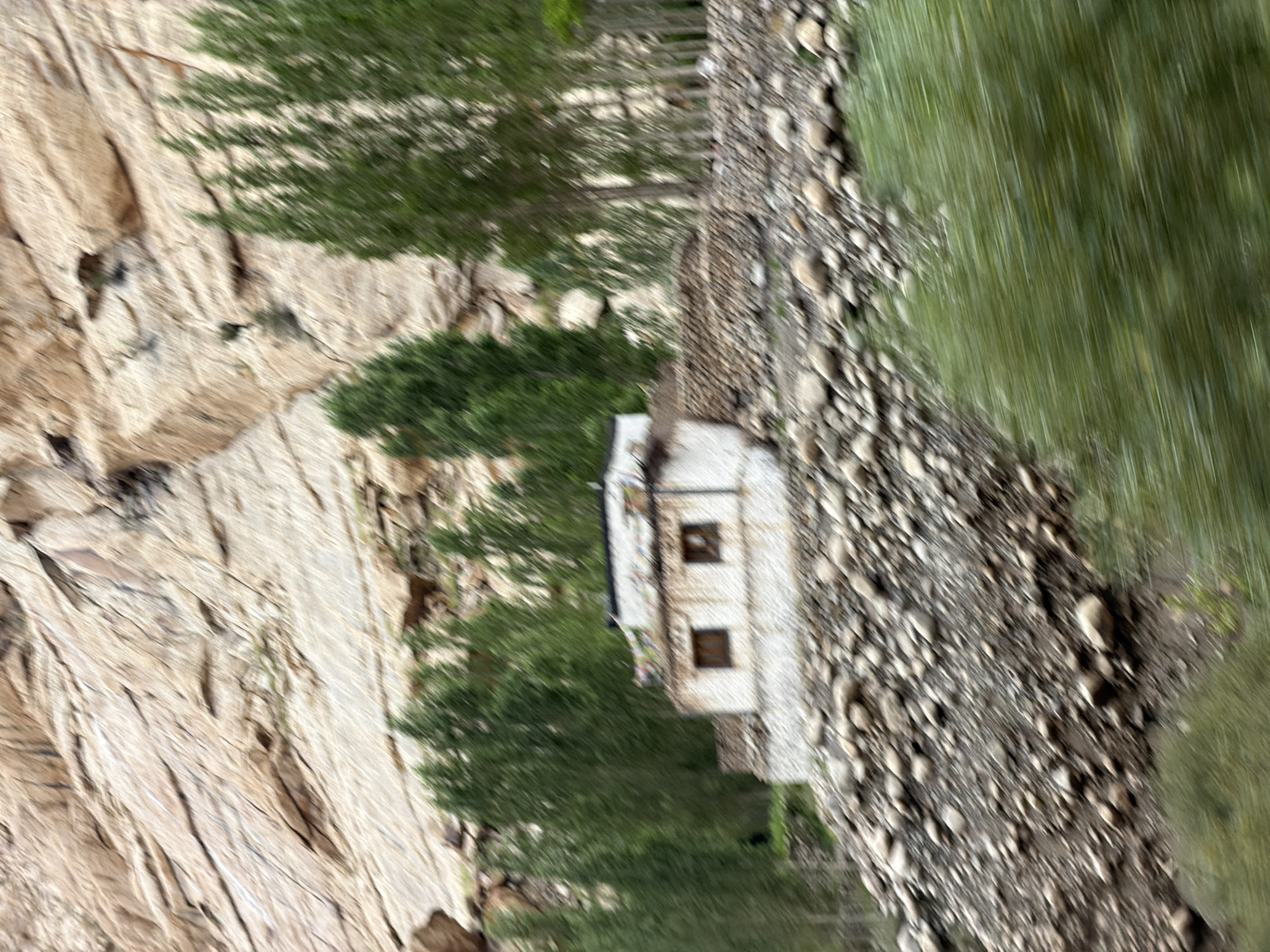 The image depicts a small, white building with two windows and a flat roof, possibly a monastery, situated on a rocky hillside in Kungyamdo, India. Prayer flags are visible on the roof. The building is surrounded by trees and a stone wall. The foreground is blurred with green foliage, suggesting movement. The background features a rocky, mountainous terrain with layered rock formations. The overall scene appears to be a rural, high-altitude environment.