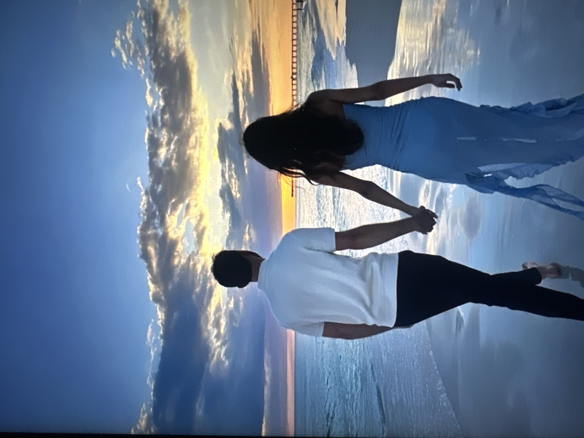 A man and a woman walk hand-in-hand along a wet sandy beach at sunset. The man is on the left, facing away from the camera, wearing a white polo shirt and dark pants. The woman is on the right, also facing away, wearing a long, strapless blue dress with ruffles. The ocean stretches out to their left, reflecting the colorful sky. In the distance, a pier extends into the water, and a bridge or structure is visible on the horizon. The sky is filled with dramatic clouds tinged with orange and yellow from the setting sun. The wet sand reflects the sky and the figures.