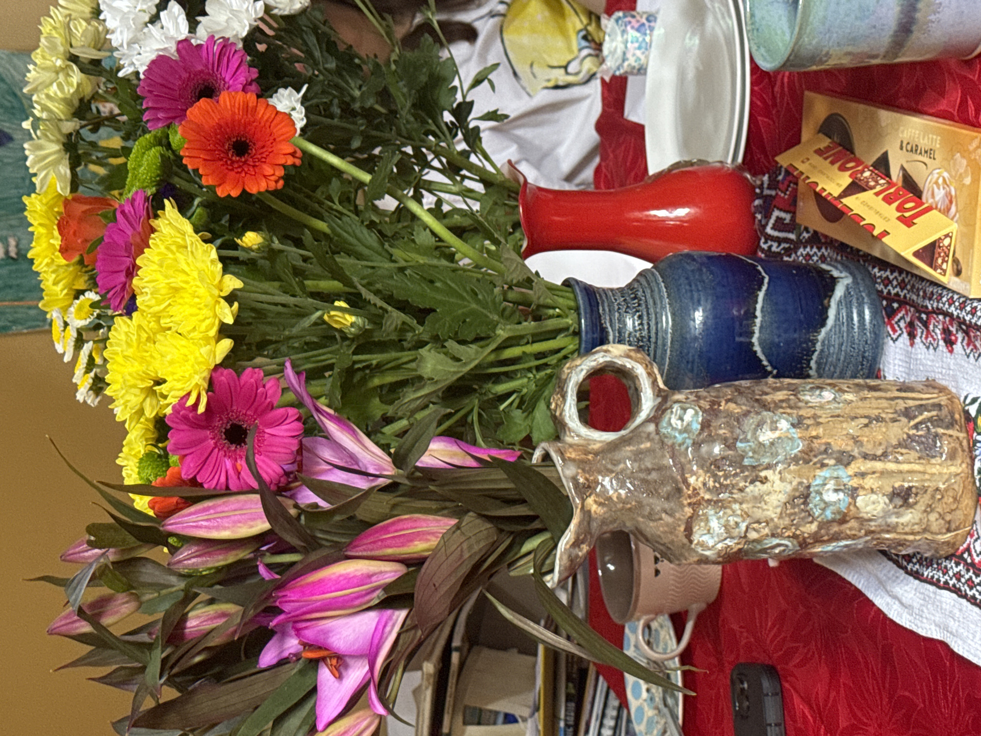 The image depicts a close-up view of a table set for a meal, adorned with a vibrant bouquet of flowers. The flowers are arranged in three vases: a rustic, earthy-toned ceramic pitcher with blue floral accents in the foreground, a dark blue glazed vase, and a bright red vase. The bouquet itself is a colorful mix of pink lilies, yellow and white chrysanthemums, and orange gerbera daisies.
On the table, beneath the flowers, are a thin yellow Toblerone chocolate bar with visible "Toblerone" text and an image of nuts and caramel, and two plates stacked one on top of the other. To the left of the vases, a light pink mug is visible, and to the far left, a dark rectangular object, possibly a mobile phone, rests on the red tablecloth.
The table is covered with a red tablecloth that has a subtle texture. A white cloth with a geometric pattern in red, black, and white is placed beneath the vases. In the background, out of focus, a person wearing a white garment with a yellow graphic can be seen. The overall lighting suggests an indoor setting, possibly a home or a restaurant. The location in the city of Waterford, Ireland, is not discernible from the visual information provided in the image.