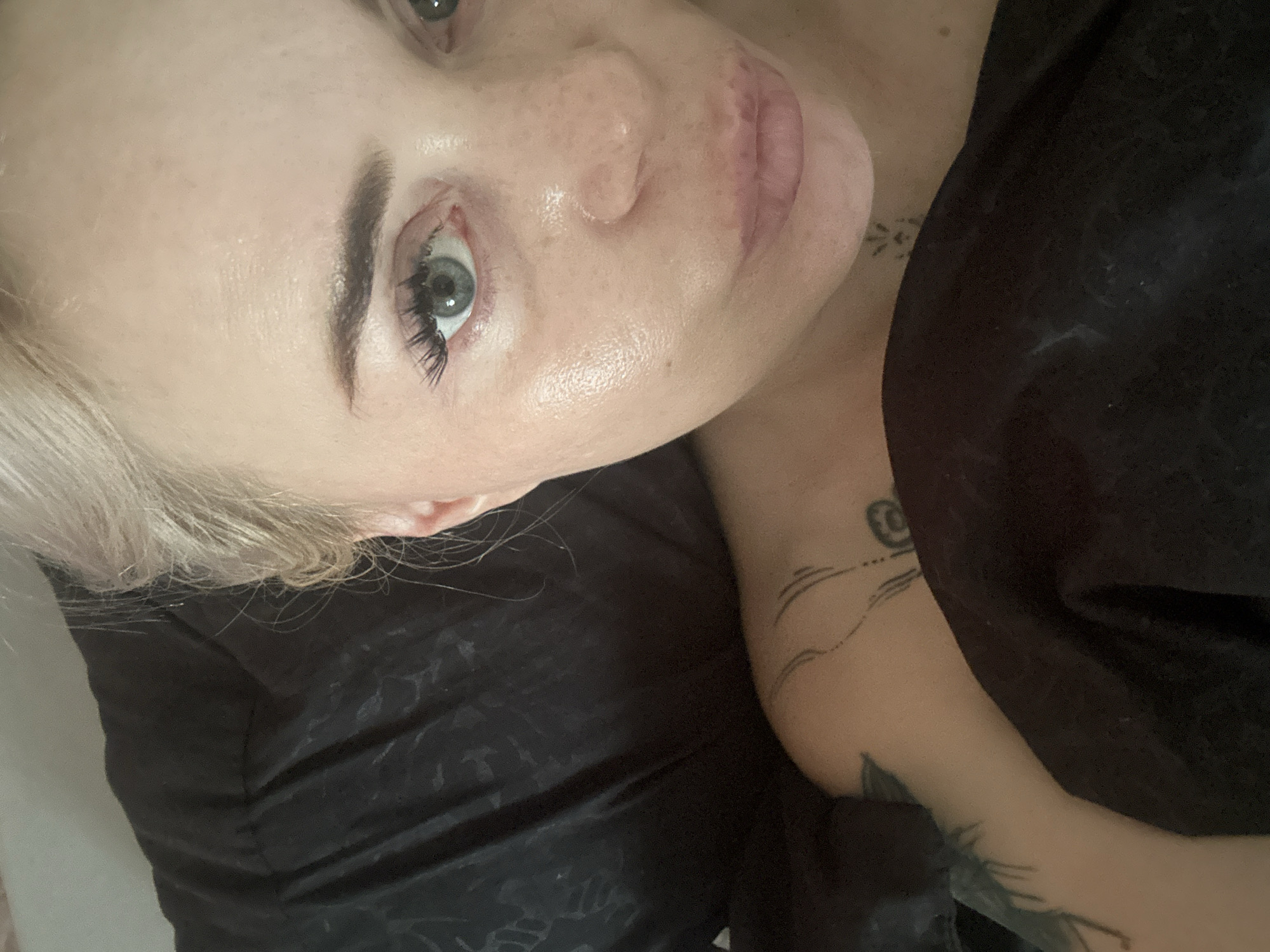 A close-up, overhead shot shows a pale-skinned woman with light blonde hair and blue eyes. She is lying down, partially covered by a dark, textured fabric that appears to be a blanket or sheet. Her face is turned towards the camera, and her lips are slightly parted. She has visible freckles on her cheeks and nose, and her skin appears slightly dewy. There are visible tattoos on her left shoulder and arm. The background is dark and out of focus. The scene appears to be indoors, possibly a bedroom.