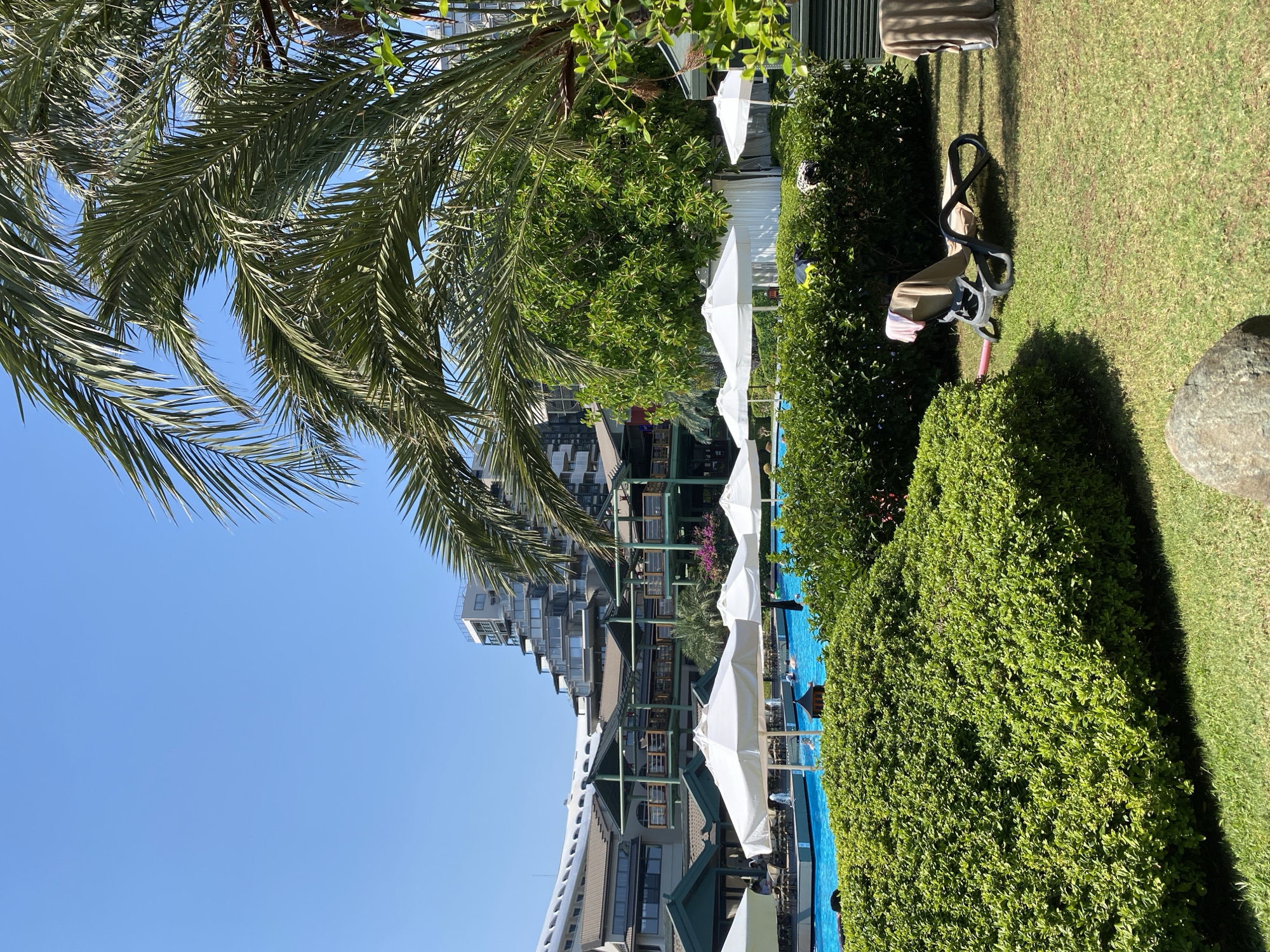 This is a medium-shot, outdoor, eye-level, vertical photograph taken in Aksu, Turkey. The scene depicts a resort pool area on a sunny day. In the foreground, a neatly manicured green lawn is met by a dense, vibrant green hedge. To the right of the hedge, a lounge chair is placed on the grass, draped with towels. Behind the hedge, a large, bright blue swimming pool is visible, dotted with several white umbrellas providing shade. A few people can be seen in the pool. In the background, a large, multi-story resort building with a unique, stepped architectural design dominates the skyline, with a clear blue sky above. Palm trees and other lush greenery frame the scene, adding to the tropical resort ambiance.