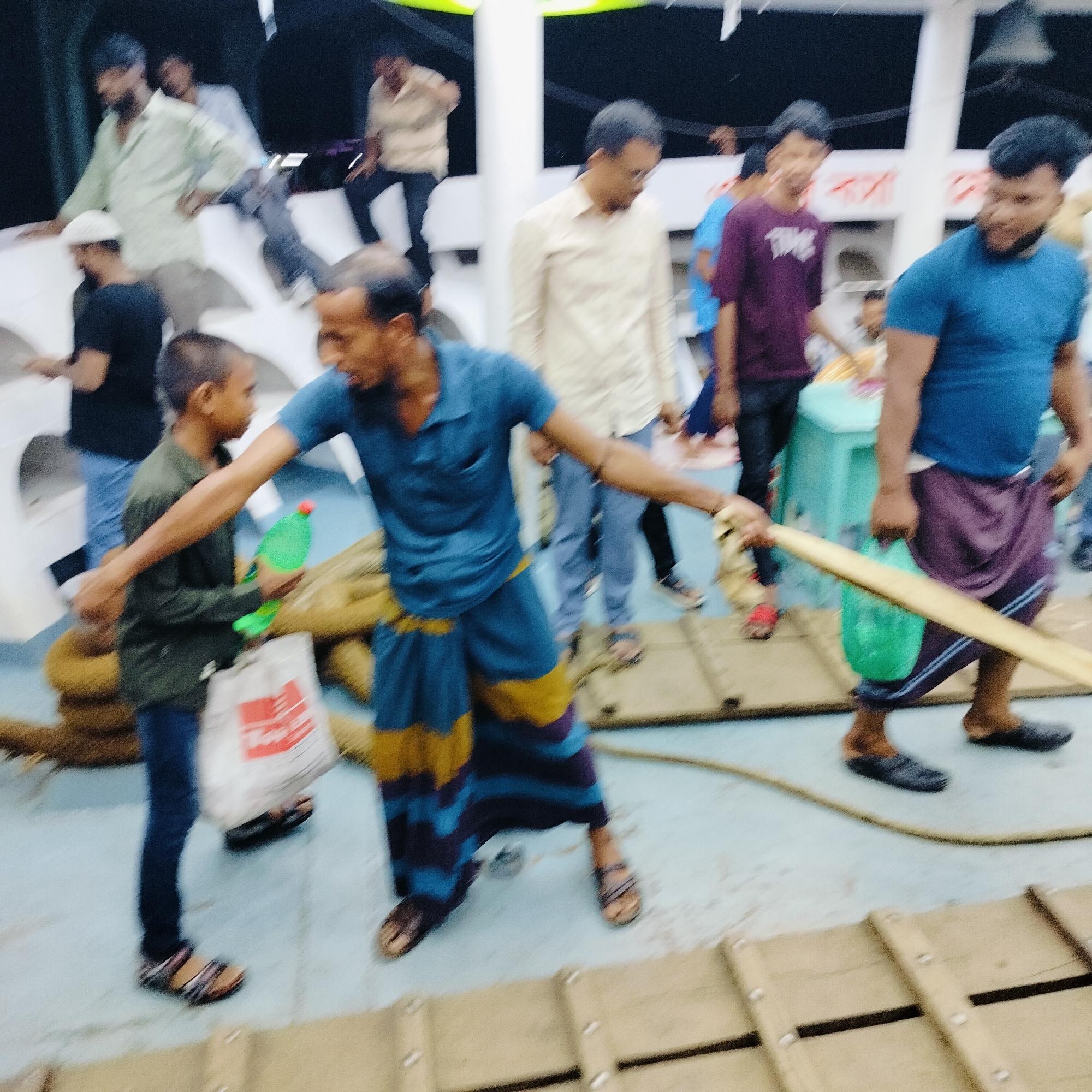 This media file captures a dynamic scene on what appears to be a crowded boat or ferry in Bhola, Bangladesh, likely during dusk or under artificial lighting. The image is characterized by motion blur, indicating movement and a fast-paced environment.
**Subjects:** The scene is populated by numerous men and at least one boy. In the foreground, a man wearing a blue polo shirt and a distinctive blue and yellow striped lungi extends his arm towards a boy. The boy, dressed in a green long-sleeved shirt and blue jeans, holds a green plastic bottle and a white bag. To the right, another man in a blue polo shirt and a purple lungi carries a green plastic bag and holds a long wooden pole. Several other men are visible in the background, some seated on boat structures, others standing, all dressed in casual attire such as shirts, pants, and lungis.
**Objects:** Prominent objects include long wooden poles, green plastic bottles, and plastic bags. Coiled ropes or woven mats are visible on the light blue deck. The floor also features wooden planks or grates. The boat's white structures and walls frame the background.
**Activity or Event:** The primary activity seems to be related to boat operations, possibly loading, unloading, boarding, or disembarking. The men handling wooden poles suggest maneuvering the vessel or cargo. The interaction between the man and boy in the foreground implies a task or exchange of items. The overall atmosphere is one of bustling activity.
**Notable Details:** The motion blur throughout the image conveys a sense of movement and energy. The attire, particularly the lungis, is consistent with Bangladeshi culture. The lighting, with darker backgrounds and diffuse light on the subjects, suggests late evening or an enclosed space.
**Visible Text:** The white plastic bag held by the boy has visible red text that appears to read "TOP". In the far background, on a white surface, there are blurry red characters that seem to be Bengali script, but they are illegible.