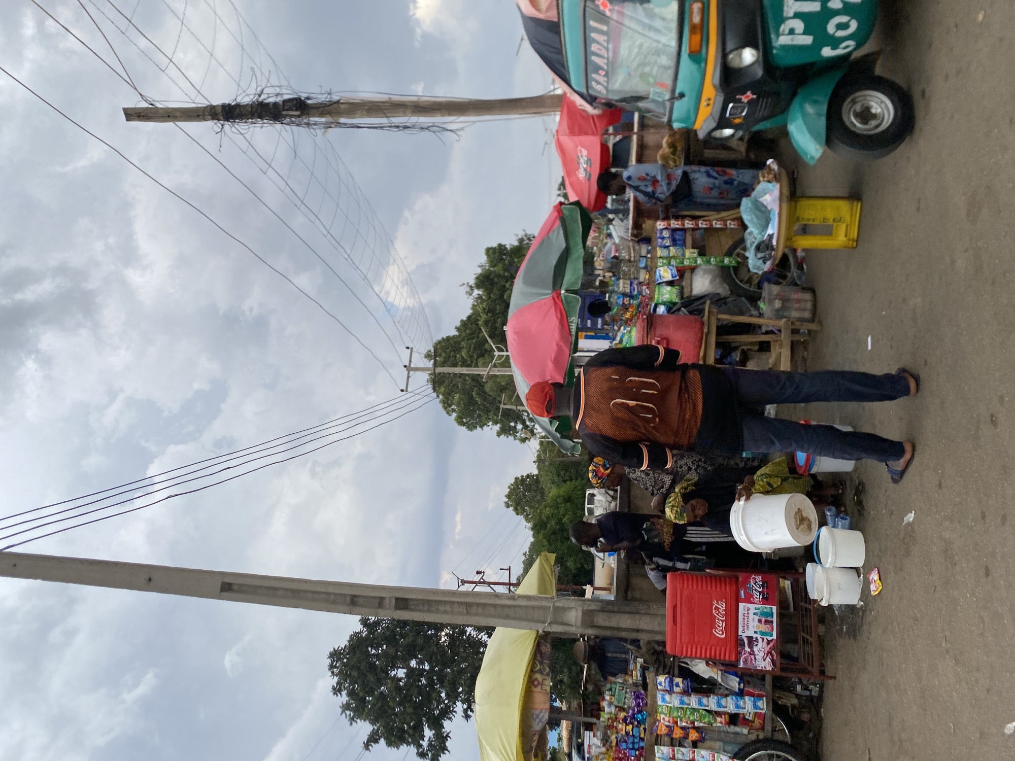 The image depicts a bustling street market scene in Jos, Nigeria. Numerous vendors display their wares on tables under colorful umbrellas and awnings, selling a variety of items, including packaged goods and snacks. Several individuals are seen browsing and interacting with the vendors. A three-wheeled vehicle, also known as a "keke," is parked on the side of the road. The street is lined with utility poles and electrical wires, and the sky above is overcast with clouds. A prominent red Coca-Cola cooler is visible, suggesting the sale of beverages.