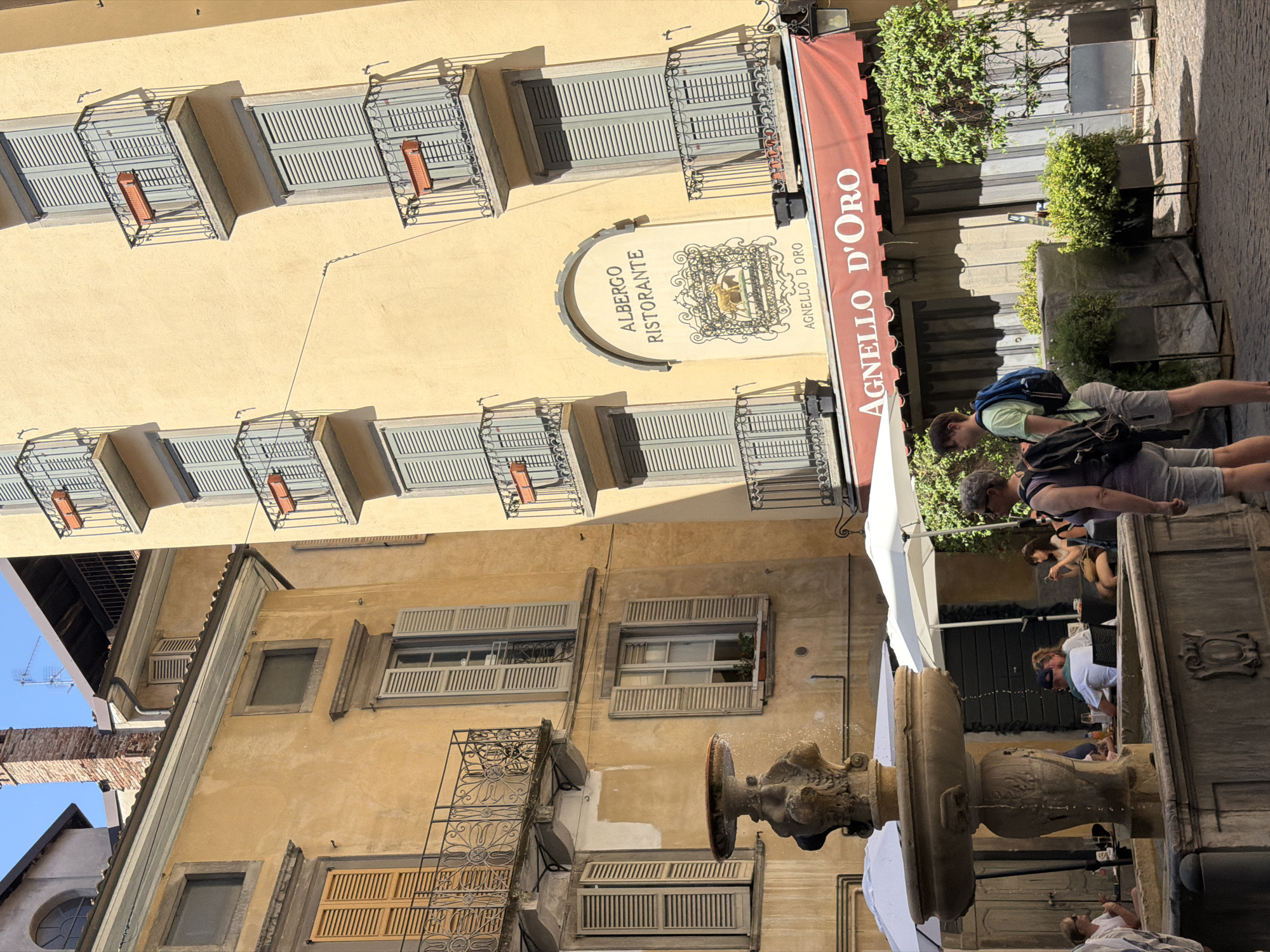 The image depicts a quaint courtyard scene in the city of Mozzo, Italy, bathed in bright sunlight. The architecture features weathered yellow stucco buildings with multiple stories, adorned with balconies featuring intricate wrought-iron railings. Several windows are visible, many with closed wooden shutters in a muted blue-gray color. A prominent feature is an antique stone fountain in the foreground, its tiered basin exhibiting intricate carvings.
In the background, an establishment named "Albergo Ristorante Agnello D'Oro" is identifiable by a sign above an arched entrance. A red awning with the same name extends over the entrance to the restaurant. The area appears to be a public space with several tables and umbrellas set up, suggesting an outdoor dining area.
Two individuals are standing in the foreground, facing away from the camera, appearing to be tourists. One person wears a light green t-shirt and shorts, carrying a blue backpack. The other wears a dark top and shorts, also with a backpack. Several other people are seated at tables in the background, some engaged in conversation, others looking at their phones. Greenery in planters adds a touch of life to the scene. The overall atmosphere is one of a relaxed European town square.