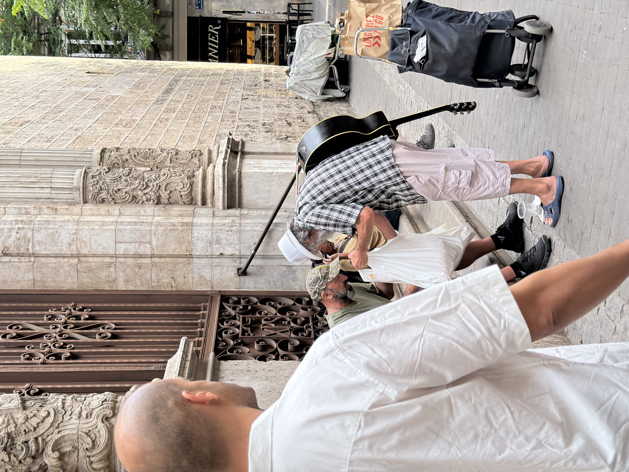 A bald man in a white shirt stands in the foreground with his back to the camera, looking towards a street scene in an urban environment. In the middle ground, two men are interacting near a decorative metal gate and a stone building with ornate architectural details. One man, wearing a camouflage baseball cap and green t-shirt, is hunched over, appearing to interact with a white plastic bag. The other man, wearing a plaid shirt and a white hat, is reaching into a white plastic bag. A black acoustic guitar rests against the plaid-shirted man's leg. The background shows a street with a bakery, a trolley, and shopping bags on the ground. The scene appears to be on a cobblestone street.