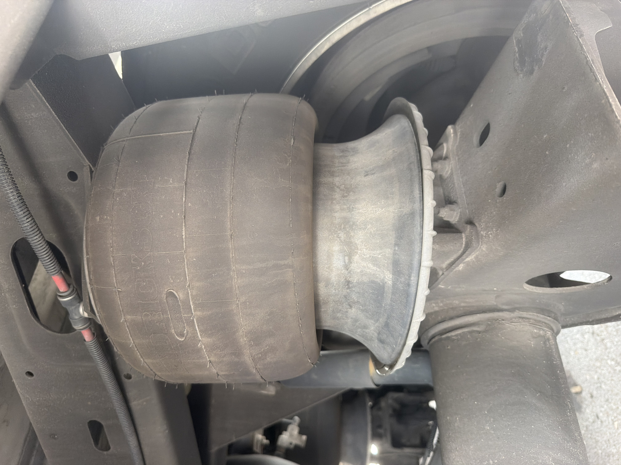 An air spring for a vehicle's suspension system is shown mounted between the frame and axle. The air spring is a black, cylindrical bellows that has text visible on its surface, which appears to read "Firestone." A thin black hose with a red wire inside is connected to the top of the air spring. The air spring is attached to a metal bracket on the vehicle's frame, and a rubber boot extends from the bottom of the air spring to a metal component of the axle. The vehicle is parked outdoors, likely on an asphalt surface, with a blurred background that suggests an urban or suburban environment. No people or specific landmarks are identifiable, and the image does not provide enough information to pinpoint the location to the city of Lebanon, United States of America.