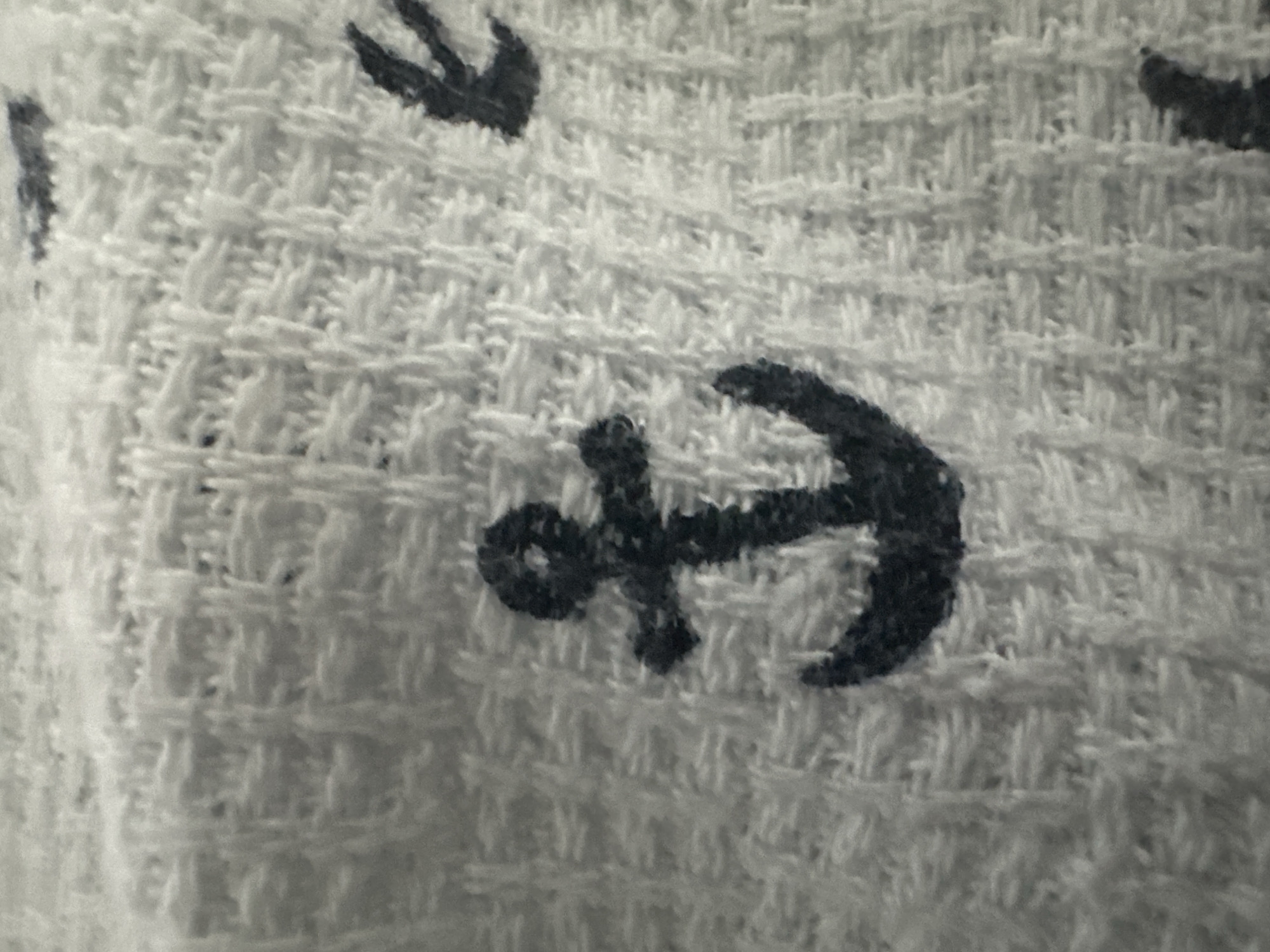 The image is a close-up, horizontal shot of a white textured fabric with dark blue anchor patterns. The texture of the fabric appears to be a waffle knit or similar weave, with raised square or rectangular sections. Several anchor motifs are visible, with the most prominent one centered in the foreground. This anchor is depicted in a dark navy blue, with a clear shank, flukes, and a crossbar. Other partial anchor shapes or similar nautical elements are visible in the background, suggesting a repeating pattern across the fabric. The lighting is even, and the focus is sharp on the fabric's texture and the printed pattern. There are no people, actions, or environmental elements from a city visible; the image is solely focused on the textile.