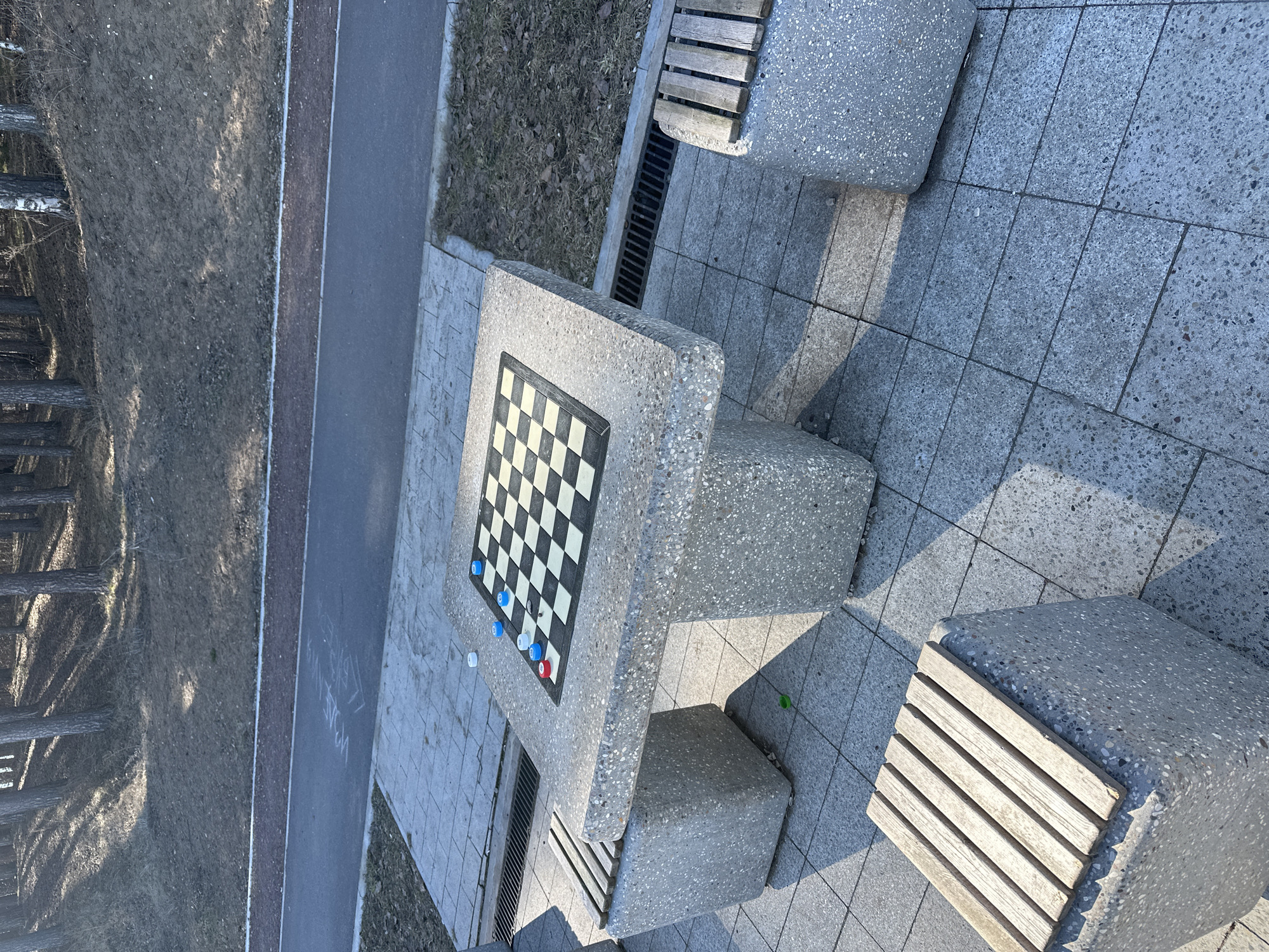 The image shows an outdoor checkerboard table with attached seating. The table and seating are constructed from a gray, speckled aggregate material. A checkerboard pattern is inlaid on the table surface, with several checker pieces scattered on the board: predominantly blue and white, with one red piece. The table is located on a paved area adjacent to a walkway. A grassed area and trees are visible in the background. The location is Chaikovsky, Russia.