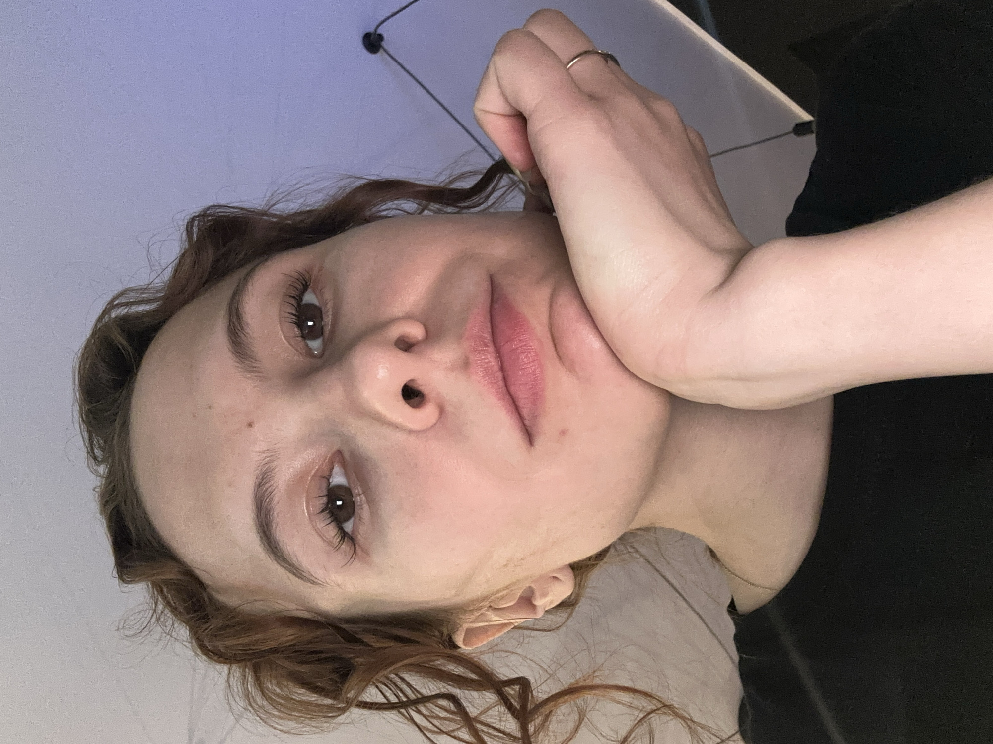 A close-up, rotated portrait captures a young person, likely female, with fair skin and wavy, light brown hair. She looks directly towards the camera with a neutral, calm expression. Her right hand, featuring a simple ring on her index finger, rests gently against her cheek and chin. Her eyes are dark brown with prominent, long eyelashes, and her lips are slightly parted, revealing a soft pink color, possibly enhanced with a light lip tint. The background consists of a plain, light purplish-blue surface, likely an indoor wall or ceiling, with darker and lighter elements visible to the right, suggesting room furniture or fixtures. The lighting is soft and even, illuminating her face. The image does not contain any discernible text or specific cues to indicate the time of day, weather, or the provided location context of Novi Sad, Serbia.