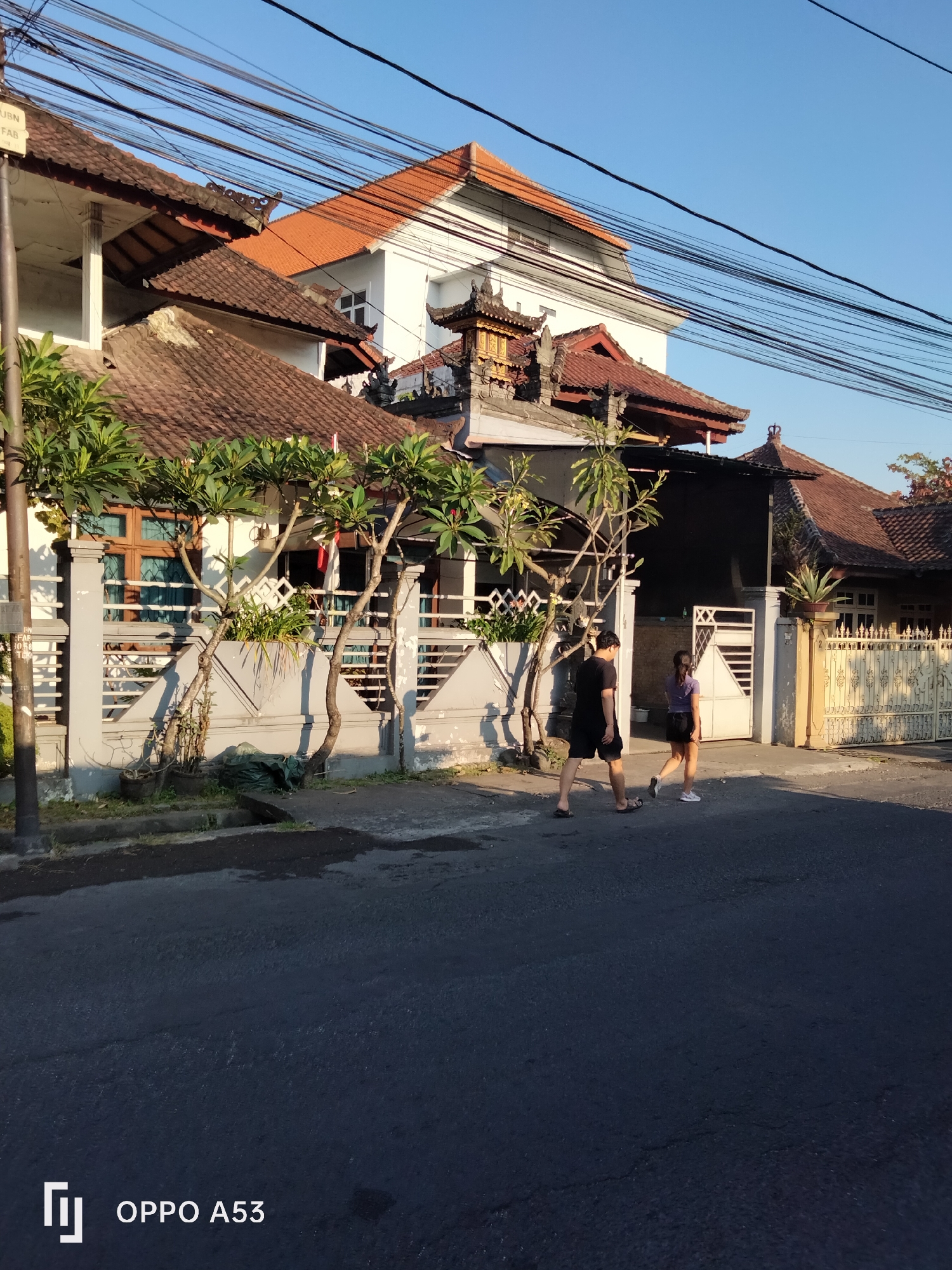 A full shot captures a street scene in Denpasar, Indonesia, with traditional Balinese architecture and a clear blue sky. The foreground is dominated by a dark asphalt road, showing some minor cracks and a slight sheen. On the left side of the road, a grey concrete fence is visible, partially obscured by potted plants and trees with green foliage. Behind the fence, a two-story building with a white facade and brown window frames is seen, topped with a brown tile roof adorned with decorative eaves. Several utility wires crisscross the sky above.
Further back, more buildings with similar brown tile roofs and white walls are present, featuring intricate architectural details reminiscent of Balinese temples. One building displays a golden decorative element on its roof.
Two individuals are walking on the sidewalk adjacent to the road. The person on the left is wearing a black t-shirt and black shorts, with dark flip-flops. The person on the right is wearing a purple t-shirt and a dark skirt, with white athletic shoes. They are walking away from the viewer and towards the right of the frame. Their interaction is neutral, as they walk side-by-side.
The overall lighting suggests it is either early morning or late afternoon, with the sun casting long shadows. The environment appears to be a residential area with a mix of modern and traditional elements.