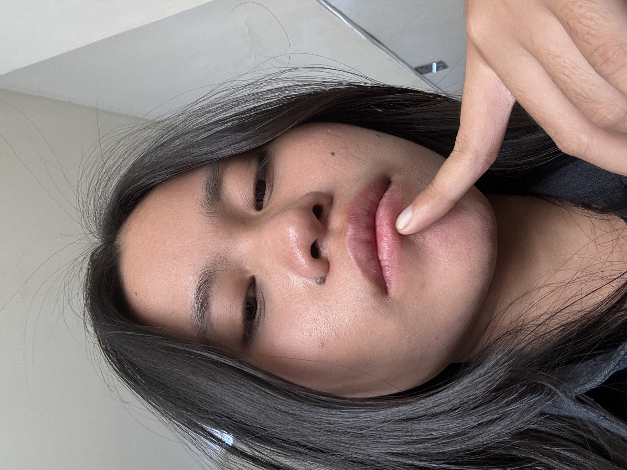 The image shows a close-up, waist-up shot of a young person with dark hair, their face taking up most of the frame. They are looking downwards, with their right index finger touching their lips. The background is blurred, but appears to be an indoor setting with a light-colored wall and a diagonal line that could be a corner or a structural element. There are some stray hairs visible around the person's face and head. The lighting is soft and diffused. The person has a small mole on their nose. No specific location in East Jakarta, Indonesia can be identified from the image.