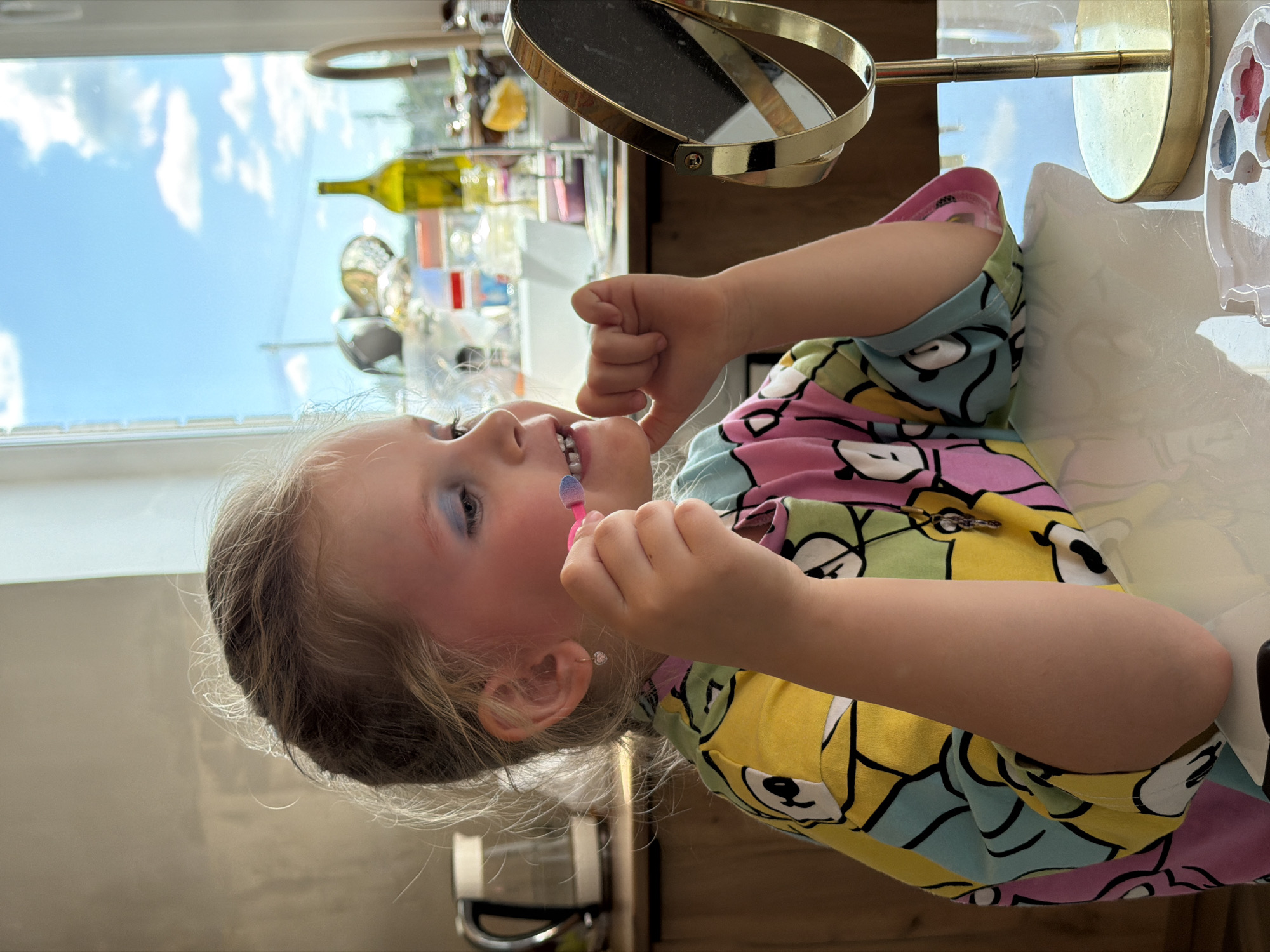 A young girl with blonde hair is applying makeup in front of a mirror, sitting at a table. She is wearing a colorful shirt with a cartoon print. Blue eyeshadow is visible on her eyelids, and she is holding a pink makeup applicator to her mouth. The background shows a kitchen area with a kettle, cabinets, and a window with a view of the sky. There is also a small makeup palette on the table in front of her. The lighting suggests it is daytime.