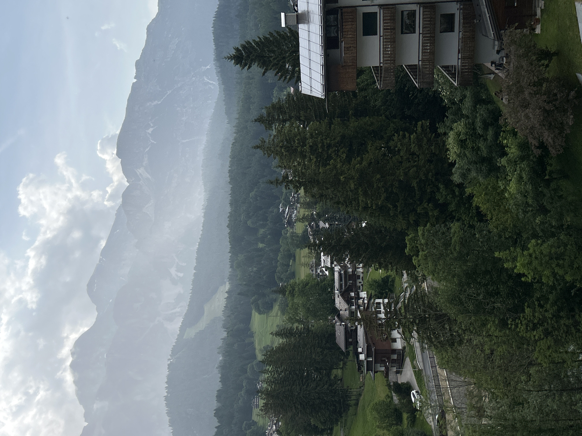 This is a vertical, eye-level, outdoor, daytime photograph of a mountain landscape with a village nestled at its base. The scene is dominated by a large, imposing mountain range that stretches across the left side of the frame. The mountains are covered in mist, obscuring their full height and detail, but their rugged, rocky slopes are visible in places.
In the foreground, on the right side of the image, is a multi-story building with a modern design featuring large windows and a solar panel array on its roof. The building appears to be a hotel or apartment complex, with balconies and outdoor seating visible. Below the building, a road winds its way through a dense forest of evergreen trees. The forest gives way to a village with several smaller buildings, mostly with dark roofs and light-colored walls, scattered amongst the trees. The village appears to be at the foot of the mountains.
The weather appears to be overcast with some breaks of sunlight creating a hazy, diffused light. The time of day is difficult to determine precisely due to the weather conditions, but it is clearly daytime. The overall mood is one of serene natural beauty, with a touch of human habitation integrated into the landscape. There is no visible text in the image.
