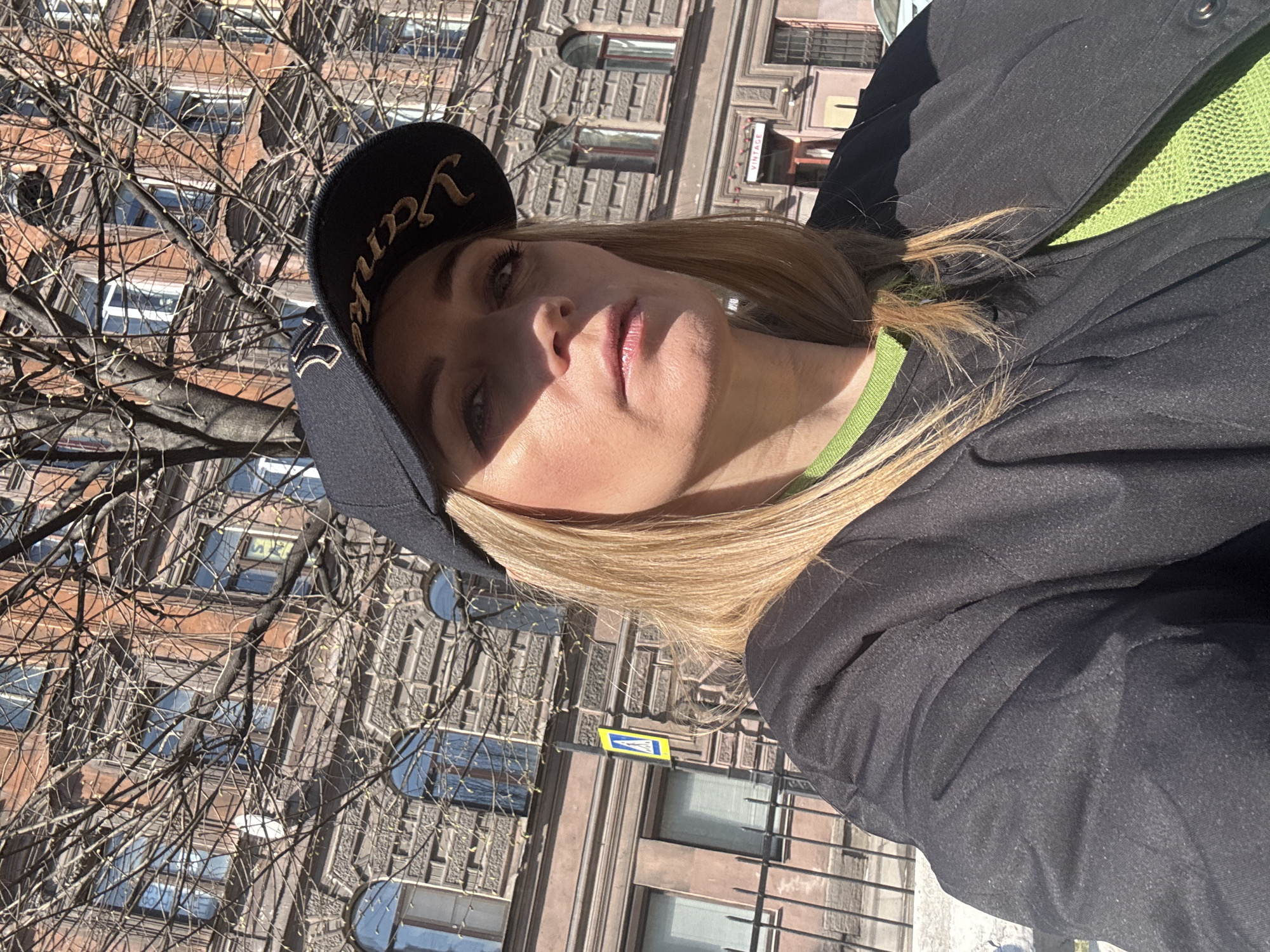 A woman poses outdoors on a sunny day. She wears a dark-colored jacket, a green shirt, and a black baseball cap with yellow embroidery. She faces the camera with her head slightly tilted to the left. Sunlight illuminates her face, casting shadows.
Behind her is a building, a tree without leaves, and a street sign. The building has multiple windows and architectural details. The street sign is blue, yellow, and white. The location is Saint Petersburg, Russia, identified by the style of architecture.
