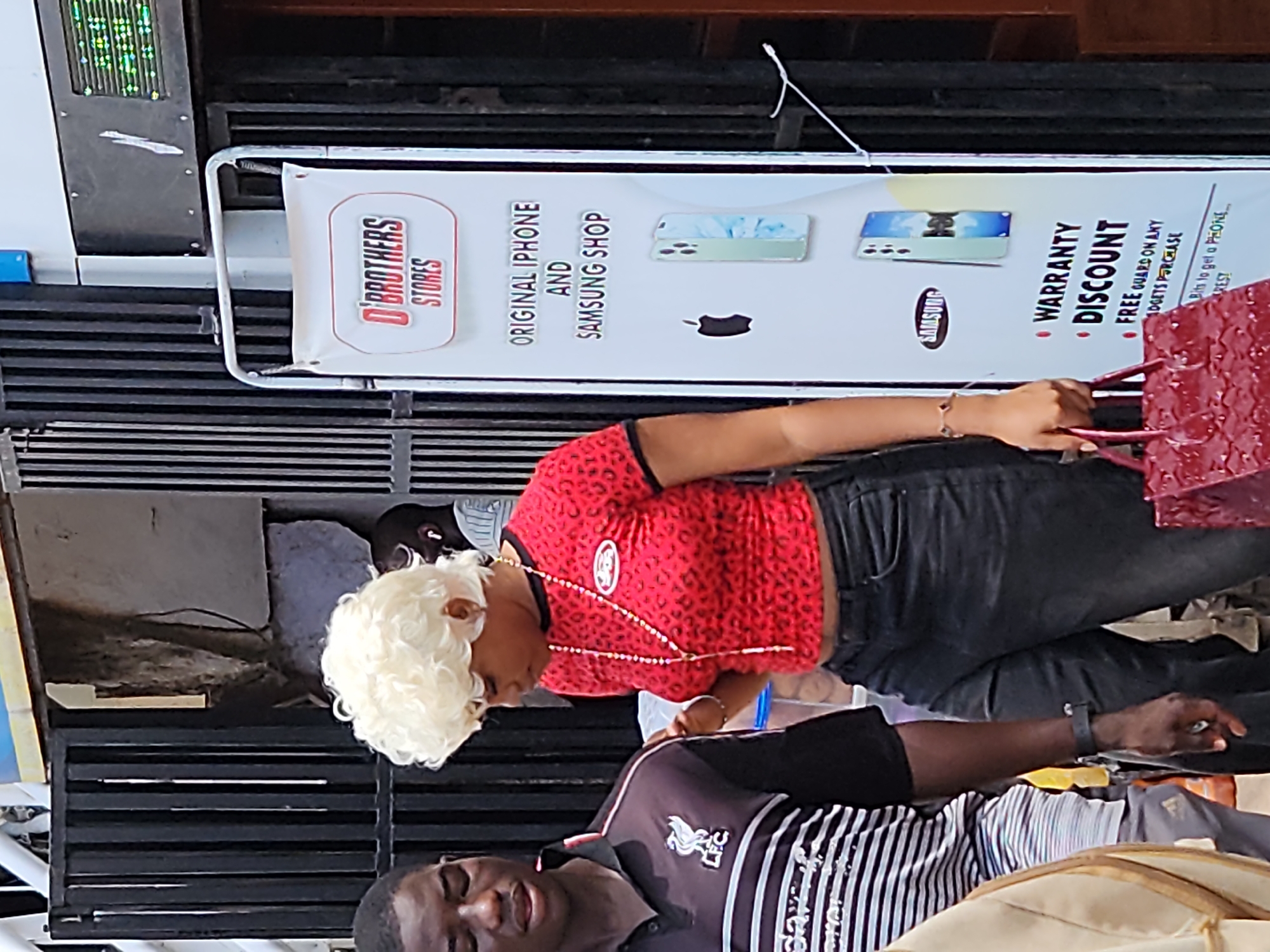 A medium shot captures a group of people standing outside a store, indicated by a large white sign with red lettering that reads "D'BROTHERS STORES". The sign advertises "ORIGINAL IPHONE AND SAMSUNG SHOP" and highlights a "WARRANTY DISCOUNT" and "FREE GUARD ON ANY GADGETS PURCHASE". The foreground is occupied by a Black male on the left, wearing a black and white striped polo shirt with the Liverpool FC logo. To his right, a Black female with blonde curly hair is seen from the chest down, wearing a red patterned top, a long beaded necklace, and dark jeans. She is holding a glossy red handbag. Behind them, another Black male is partially visible, wearing a light-colored shirt and earpods. The background features a black metal gate and a rough-textured wall, suggesting an urban outdoor setting.