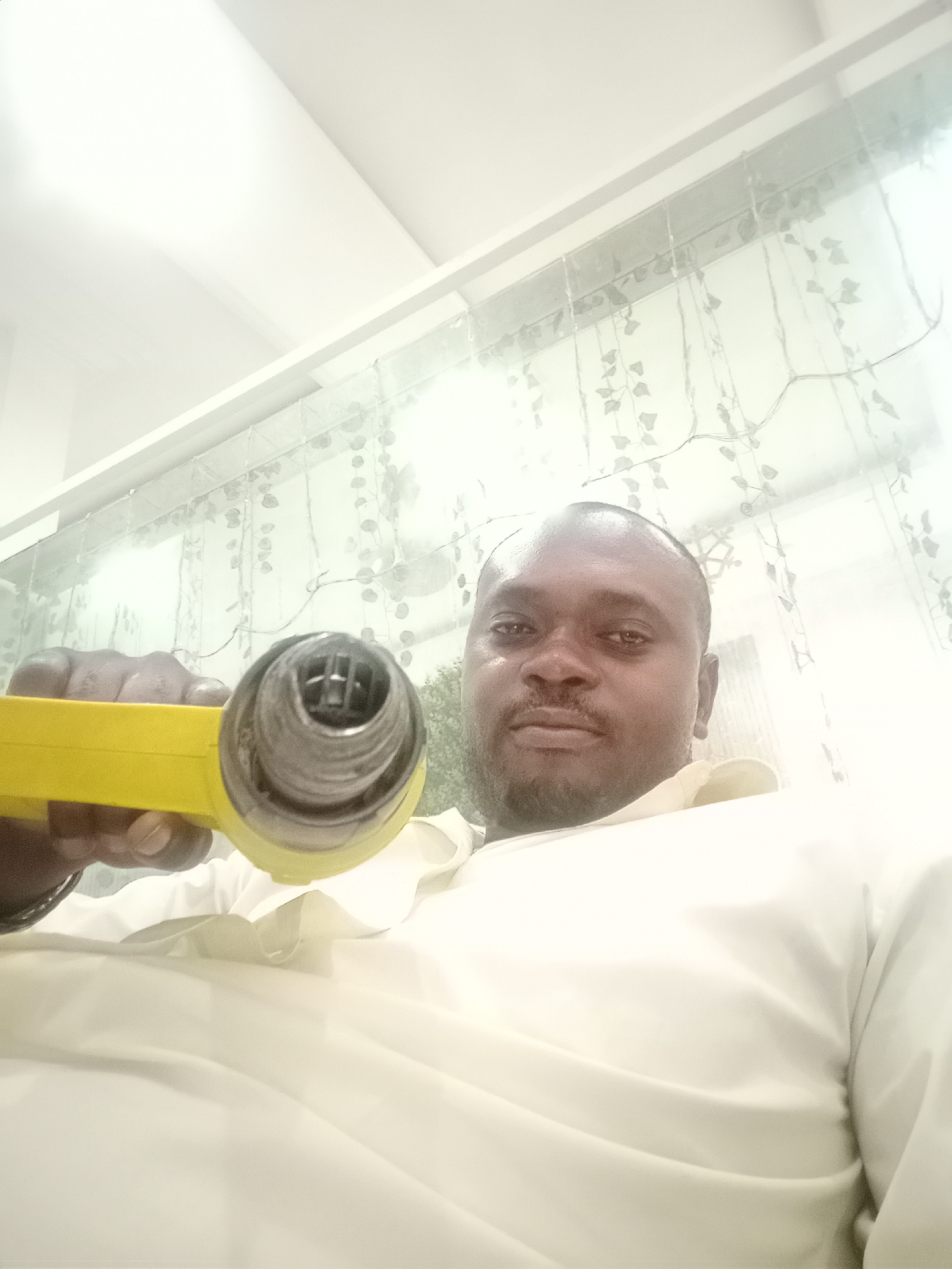 The image shows a man holding a yellow heat gun. The man is dark-skinned with a trimmed beard and is wearing a white shirt. The background includes a white ceiling and a translucent curtain with leaf-like patterns. The photograph appears to have been taken indoors. The location is Gwarinpa, Nigeria.