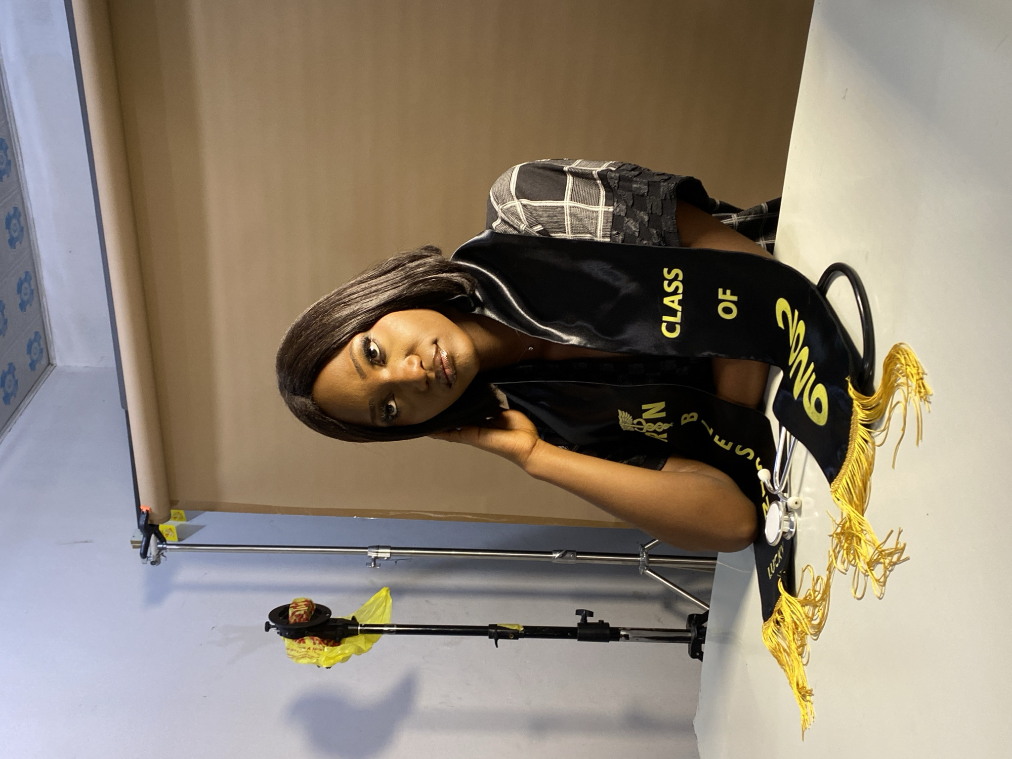 A woman is posing for a graduation portrait indoors. She is wearing a black graduation stole that says "CLASS OF 2016" and has yellow tassels. She has a stethoscope hanging from the stole. She is wearing a checkered top. The backdrop is a tan-colored photography backdrop. There is photography equipment visible in the background, including stands and clamps. The walls are painted a light blue color. It cannot be determined if this is in Gura Top, Nigeria.