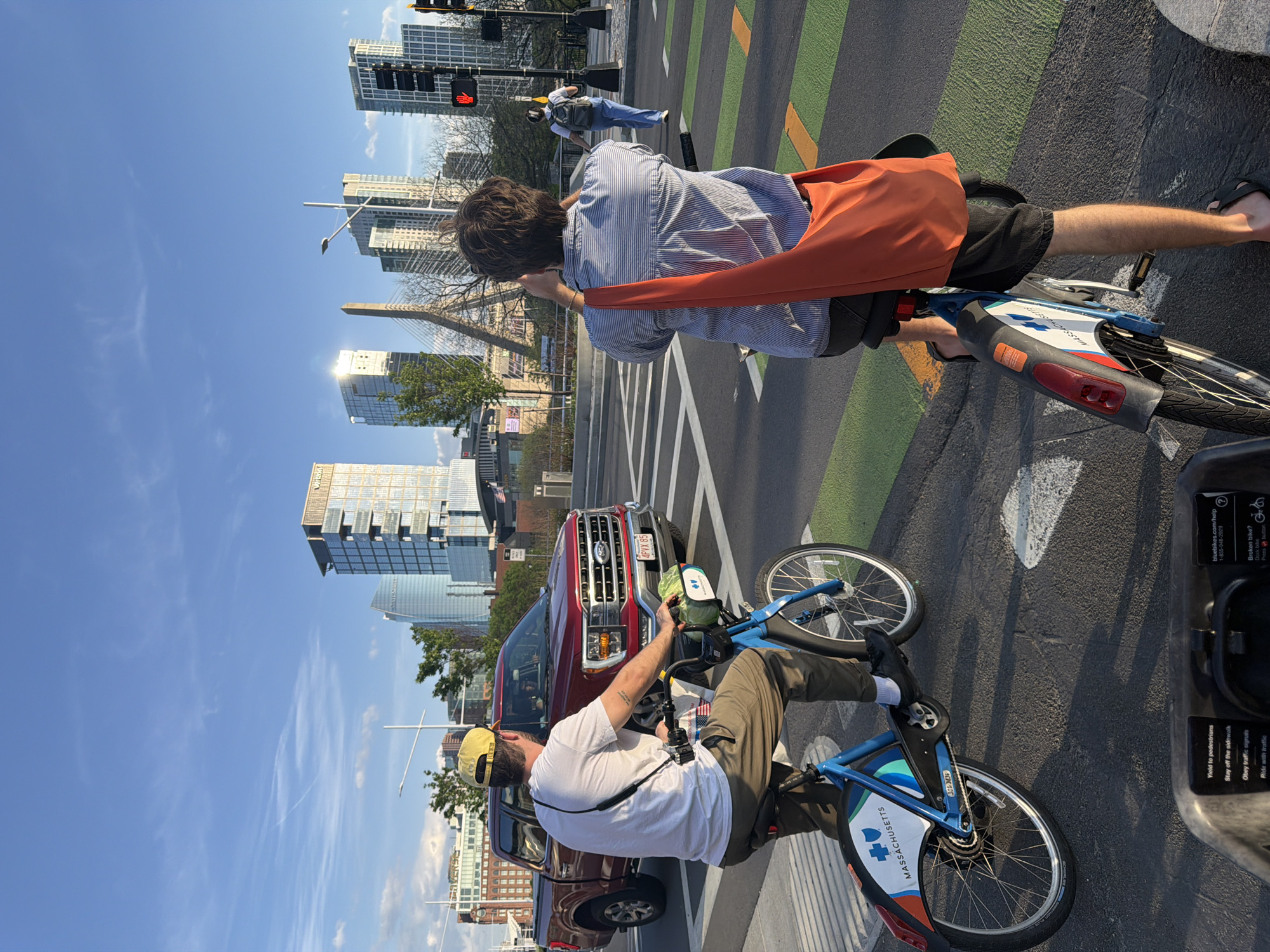 Here is a description of the image:
The image shows two people renting and riding bicycles at a Bluebikes station in Boston, Massachusetts. The bicycles are blue and have the Bluebikes logo. The bikes have a sticker that reads "MASSACHUSETTS" and "bluebikes.com/help" with a phone number.
One person is wearing a blue and white striped shirt, black shorts, and sandals and has an orange bag across their body. The other person is wearing a white t-shirt, khaki pants, and a yellow hat. The second person also has a camera around his neck.
In the background, there is a red Ford F150 pickup truck with the license plate "2PY95". There are a number of buildings in the background, with the Zakim Bridge. A traffic light is visible, showing a red light. Pedestrians are walking in the crosswalk. The sky is blue with some clouds.