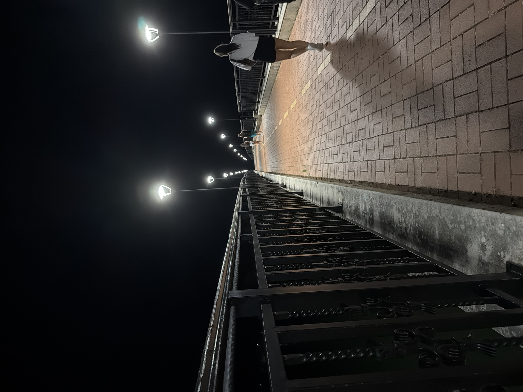 A woman with dark hair pulled into a ponytail walks away from the camera on a paved pedestrian walkway at night. She is wearing a white t-shirt and black shorts. The walkway is illuminated by streetlights spaced along the side of the path. A black metal railing runs along the left side of the walkway, separating it from a dark expanse. In the distance, two other figures are walking on the same path, also illuminated by the streetlights. The pathway is made of red bricks laid in a rectangular pattern, with a yellow line marking the center. The scene appears to be a promenade or bridge in the city of Pravets, Bulgaria.